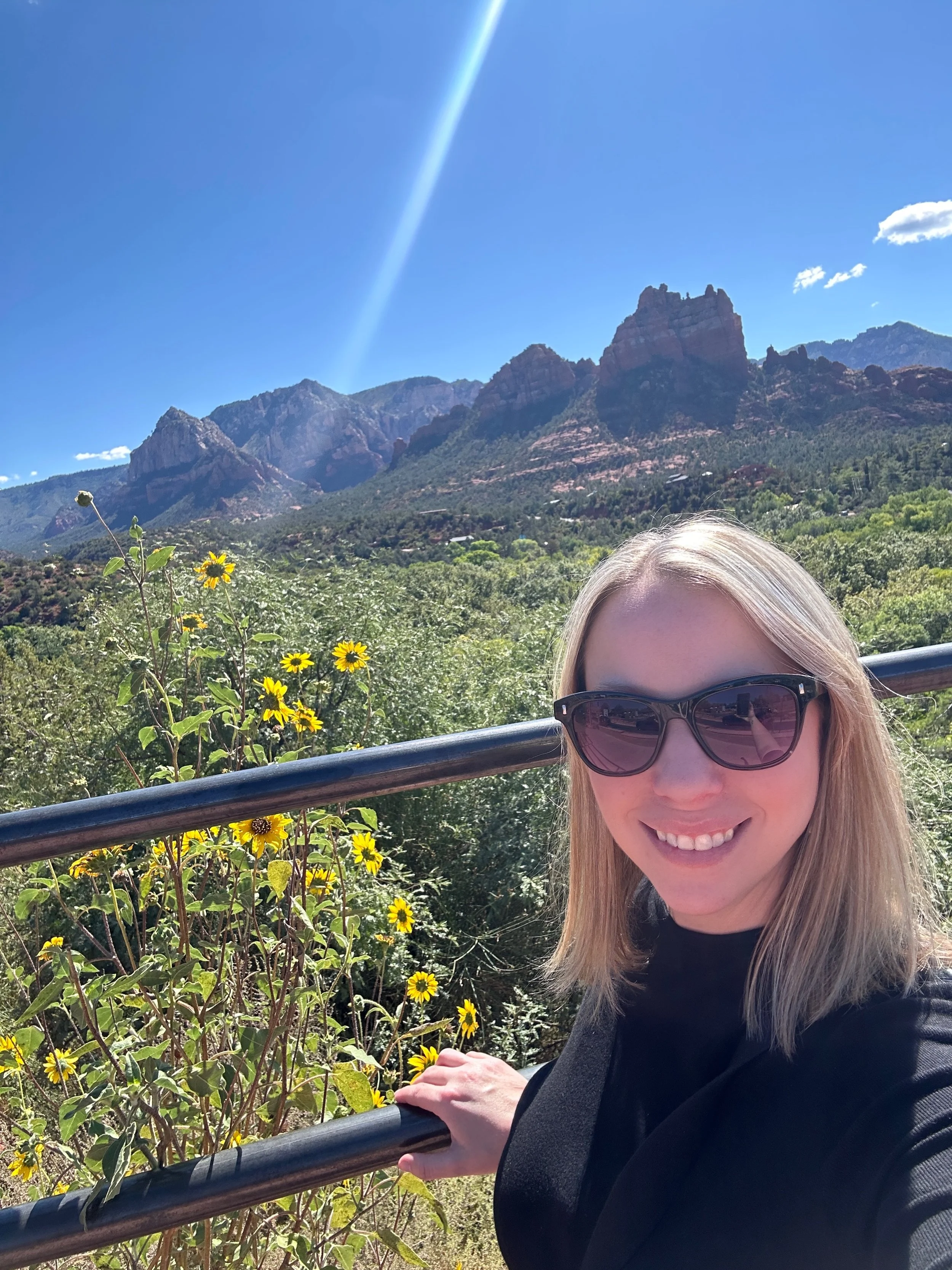 Woman in black shirt and sunglasses posing against a railing with yellow flowers and large rock formations in the background with bright blue sky and sun flare