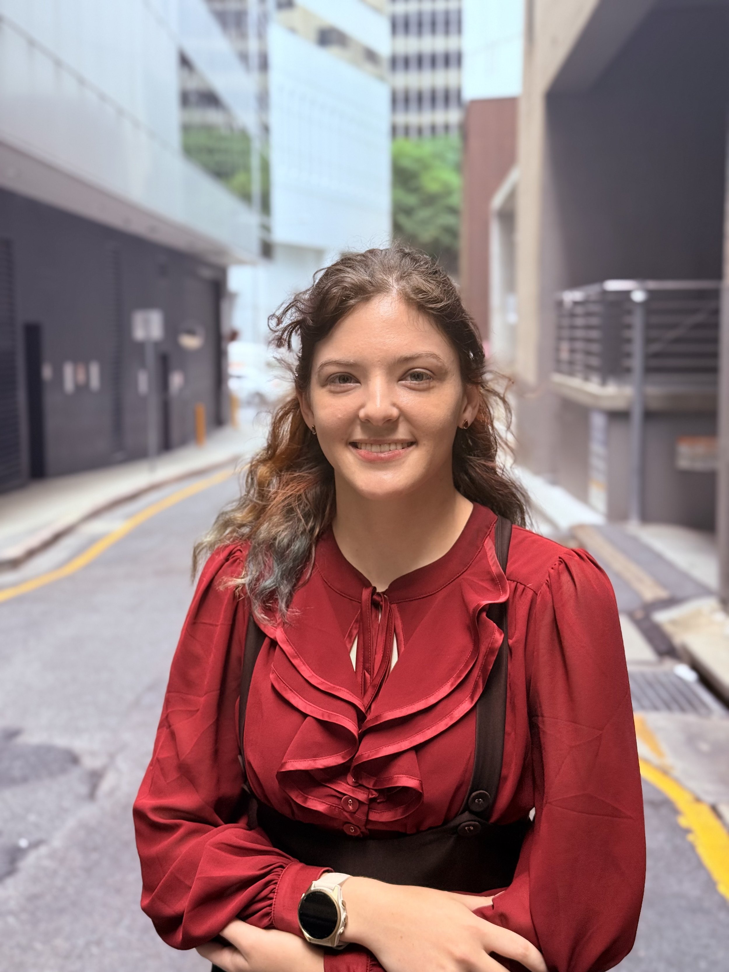 A woman with curly brown hair, wearing a red blouse with ruffles and a black vest, standing on a city street with modern buildings in the background, smiling at the camera.