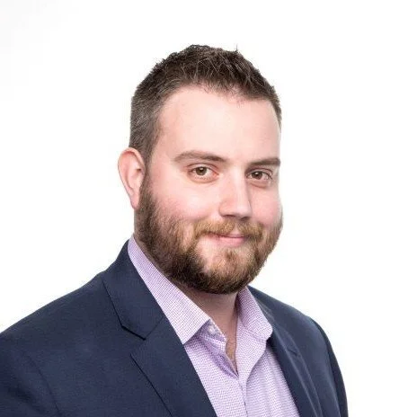 A man with short brown hair, a beard, and light skin, wearing a navy blazer and a light purple dress shirt, smiling at the camera against a white background.