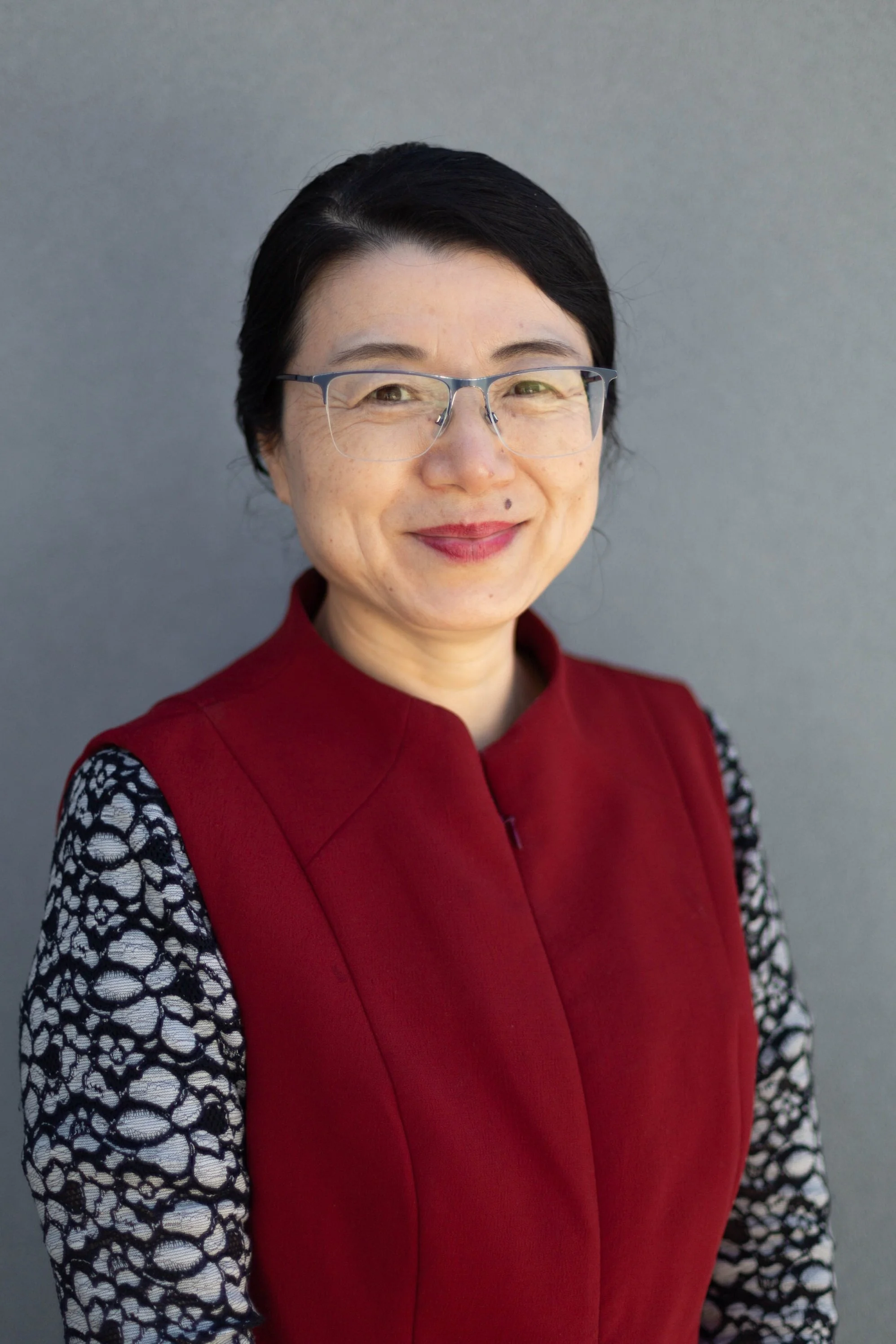 Portrait of a woman with short black hair, glasses, wearing a red vest over a patterned black and white top, smiling against a light gray background.