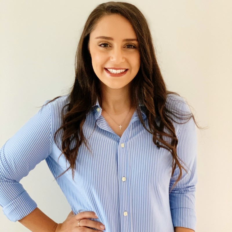 A young woman with long, wavy brown hair smiling and wearing a blue and white striped button-up shirt with a cross necklace, posing against a plain white wall.