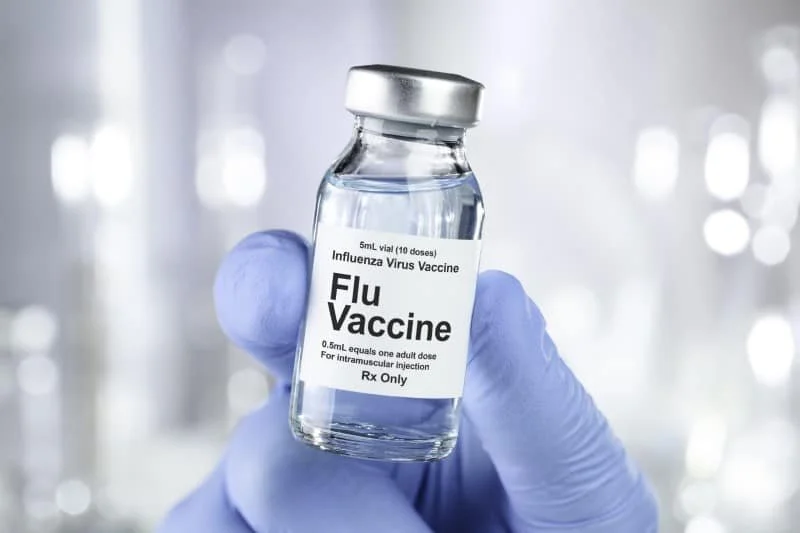 A hand wearing a blue glove holding a small glass vial labeled 'Flu Vaccine' and 'Influenza Virus Vaccine' in a clinical setting.