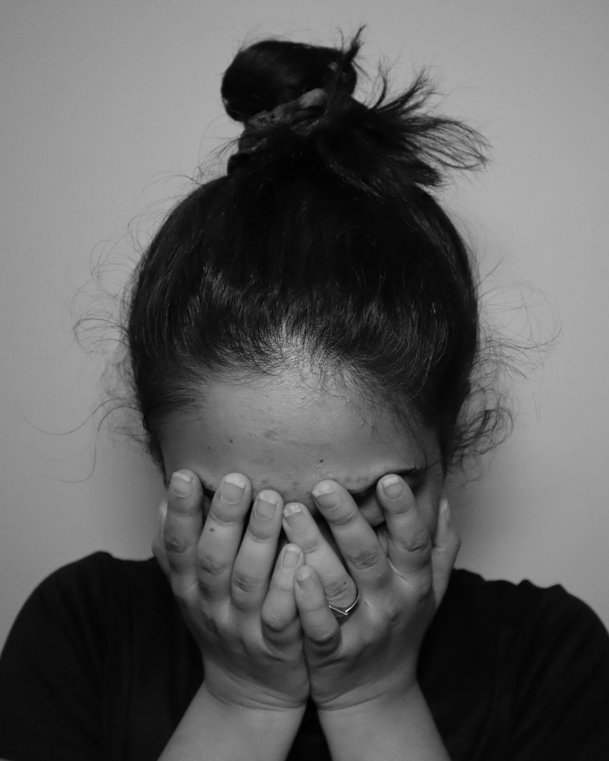 A person with dark hair styled in a messy bun covering their face with their hands.