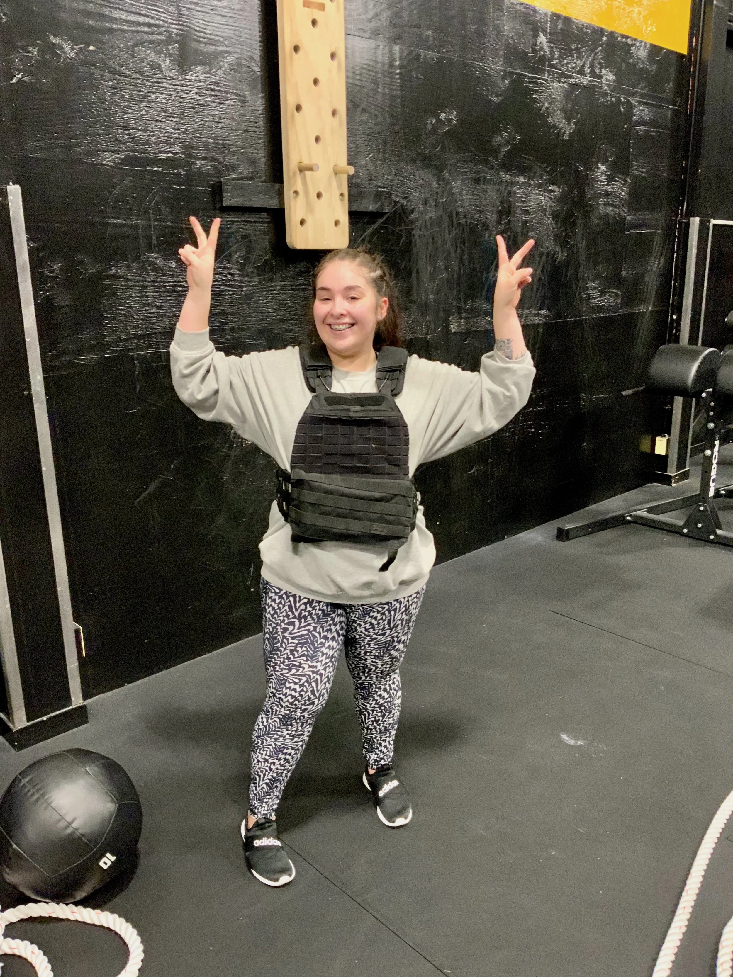A smiling woman wearing a beige sweatshirt, black tactical vest, patterned leggings, and black sneakers standing in a gym, making peace signs with both hands.