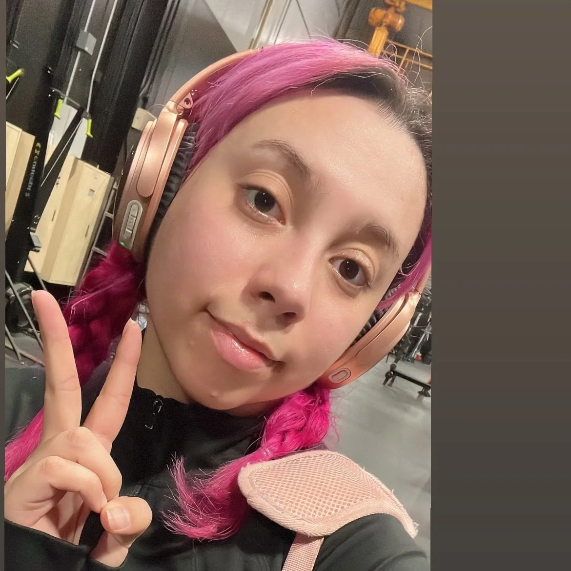 Young woman with pink hair and headphones making a peace sign in a gym or fitness studio.