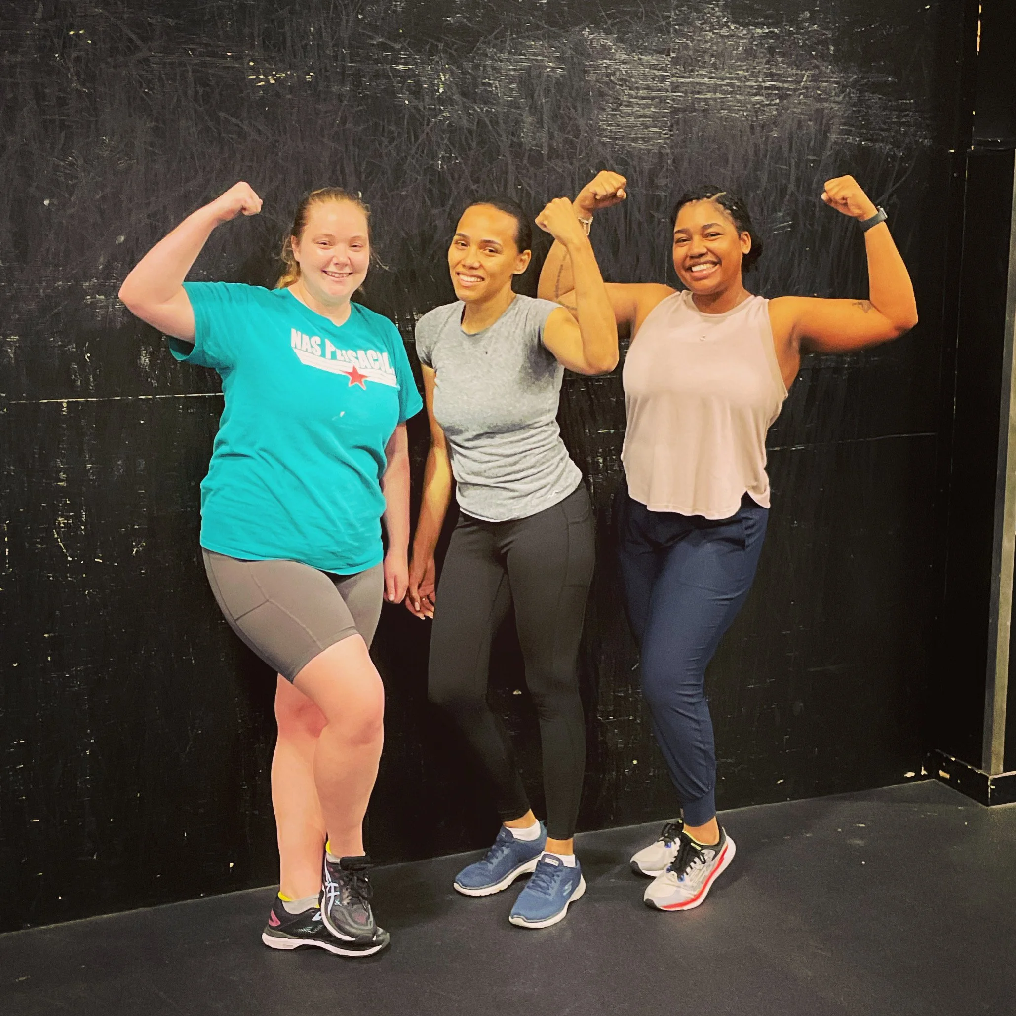 Three Military Spouse women flexing their arm muscles and smiling in a gym or fitness studio.
