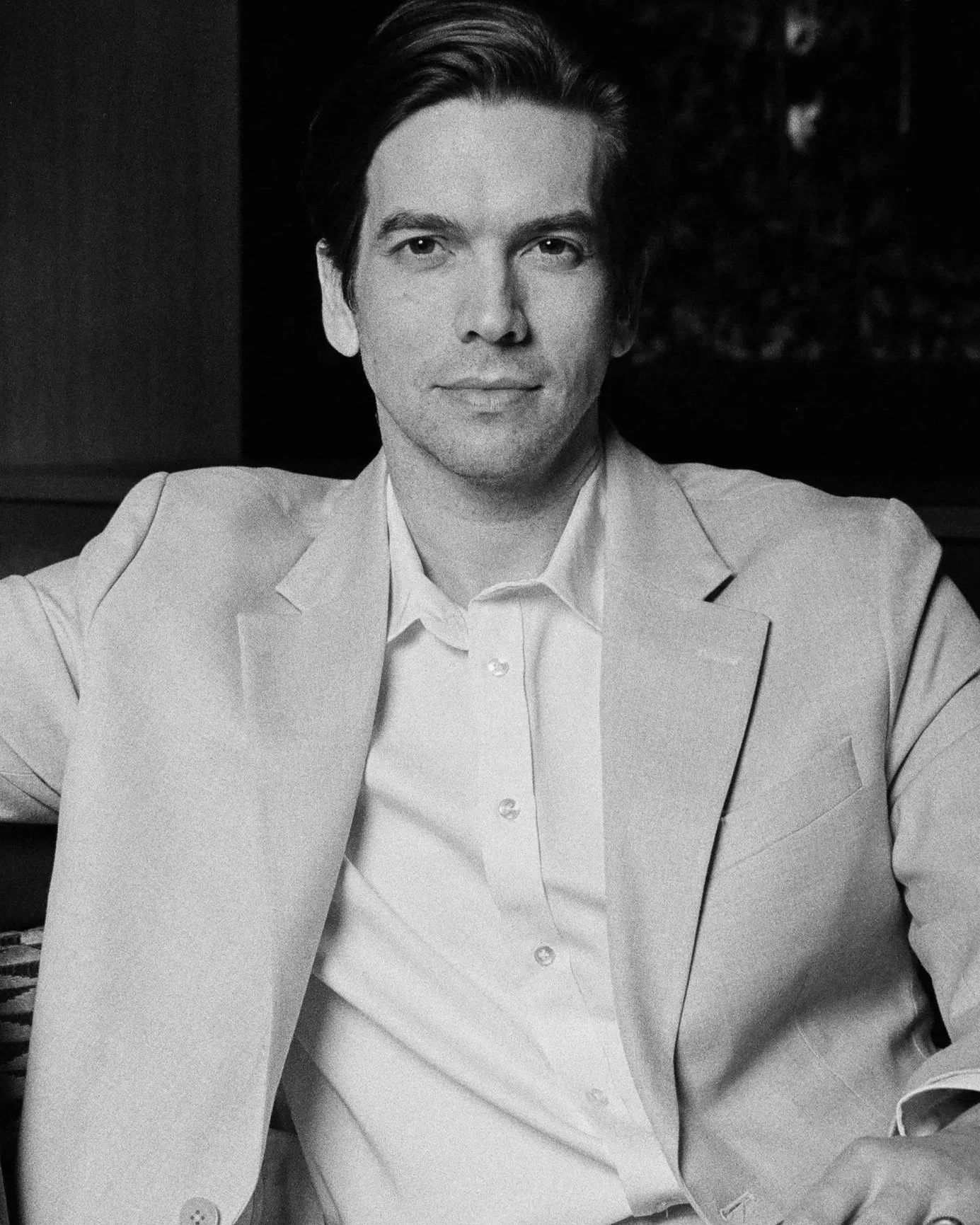 Black and white portrait of a man wearing a suit jacket and collared shirt, sitting with arms relaxed on a surface, looking straight at the camera with a slight smile.