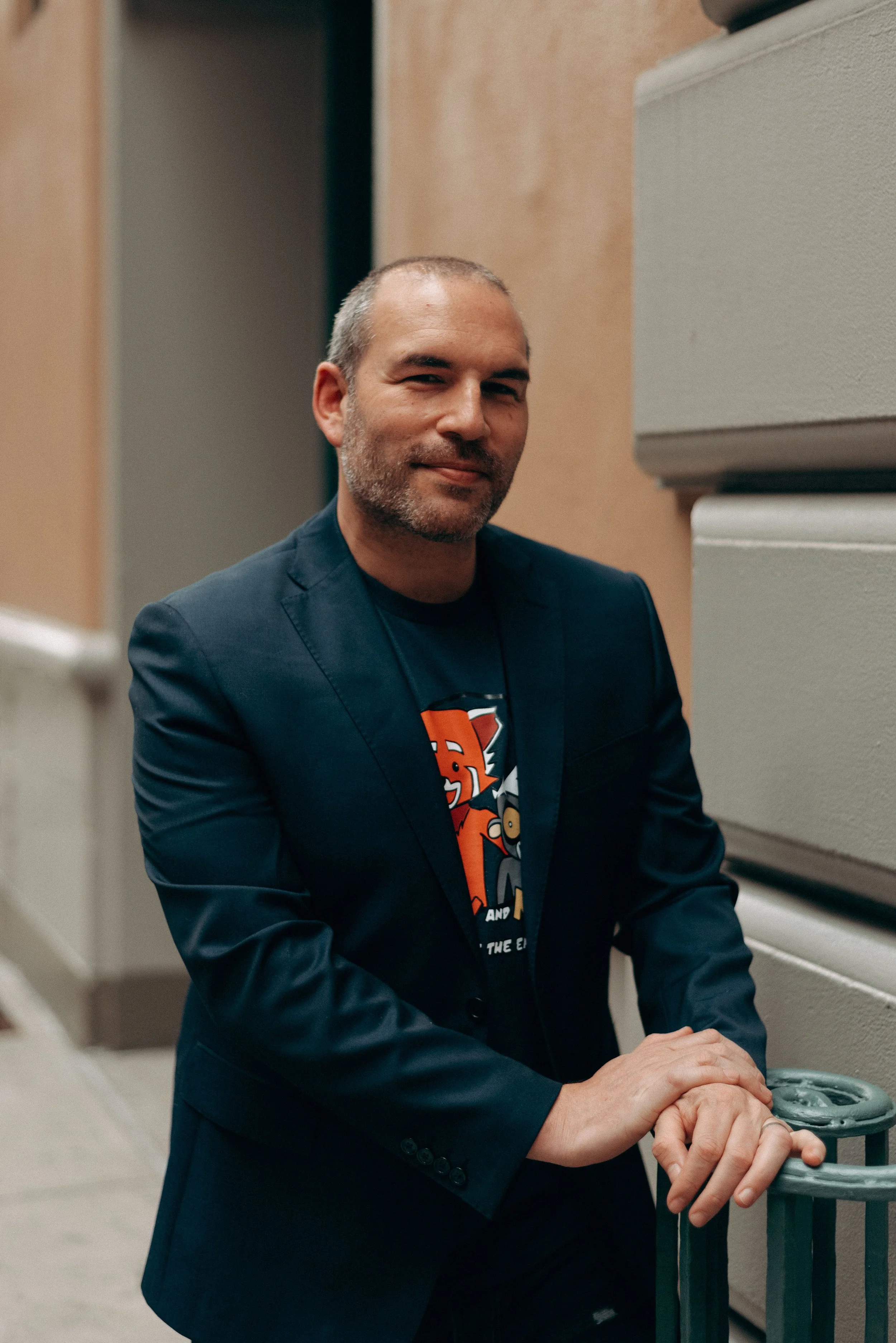 A man with short gray hair and a beard, wearing a dark blue blazer and a graphic T-shirt, is seated at a railing with his hands resting on it, smiling slightly at the camera.