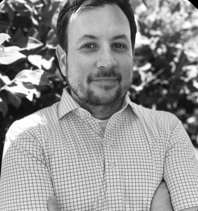 A man with short dark hair and facial hair standing outdoors in front of leafy plants, wearing a checkered collared shirt, smiling softly.