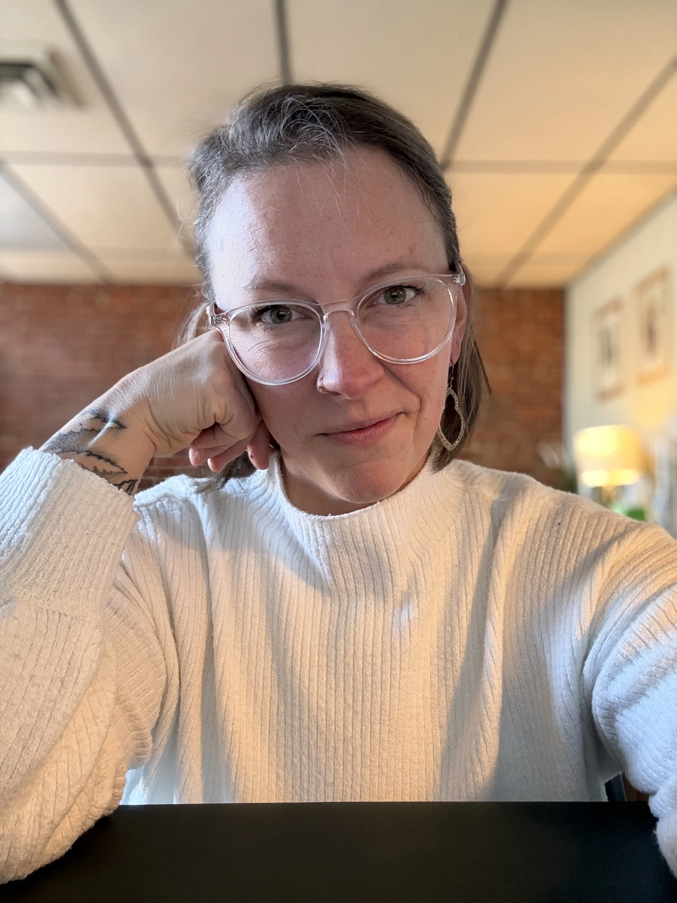 A woman with glasses smiling in a cozy living room with brick wall and framed pictures in the background.