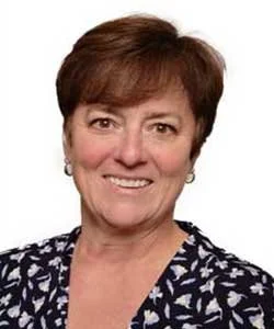Smiling person with short brown hair, wearing a dark patterned shirt, against a white background.