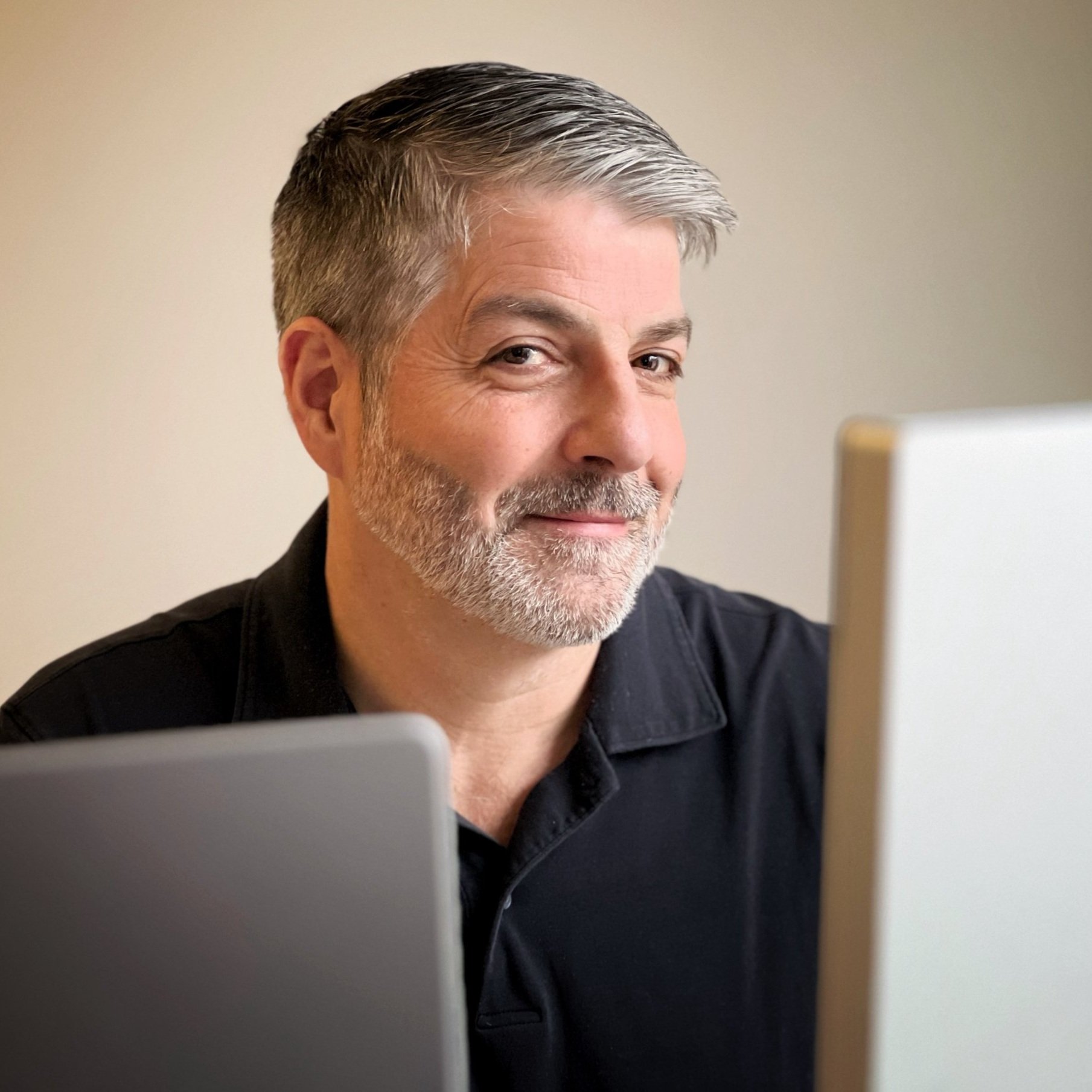 A man with gray hair and a beard smiling, seated between two computers, wearing a dark shirt.