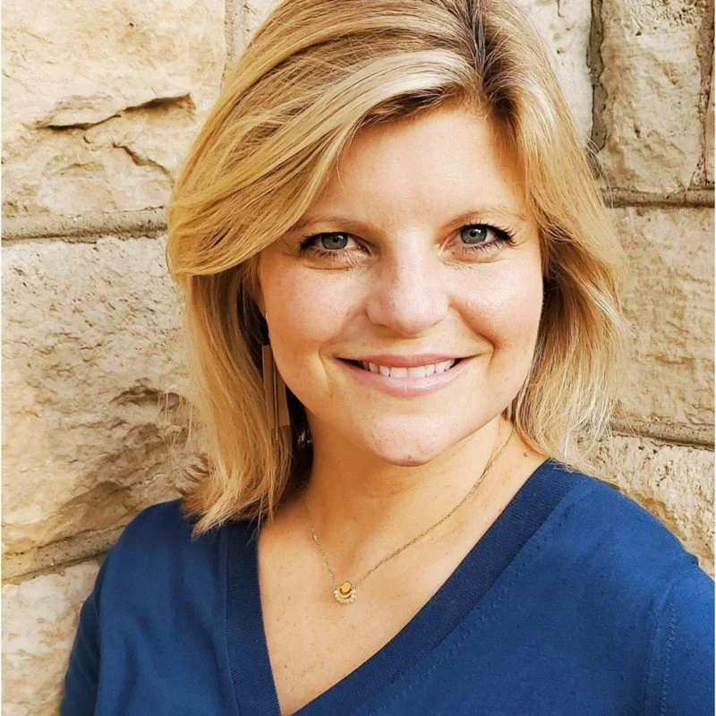 Smiling woman with blonde hair, wearing a blue top and a necklace, standing in front of a stone wall.
