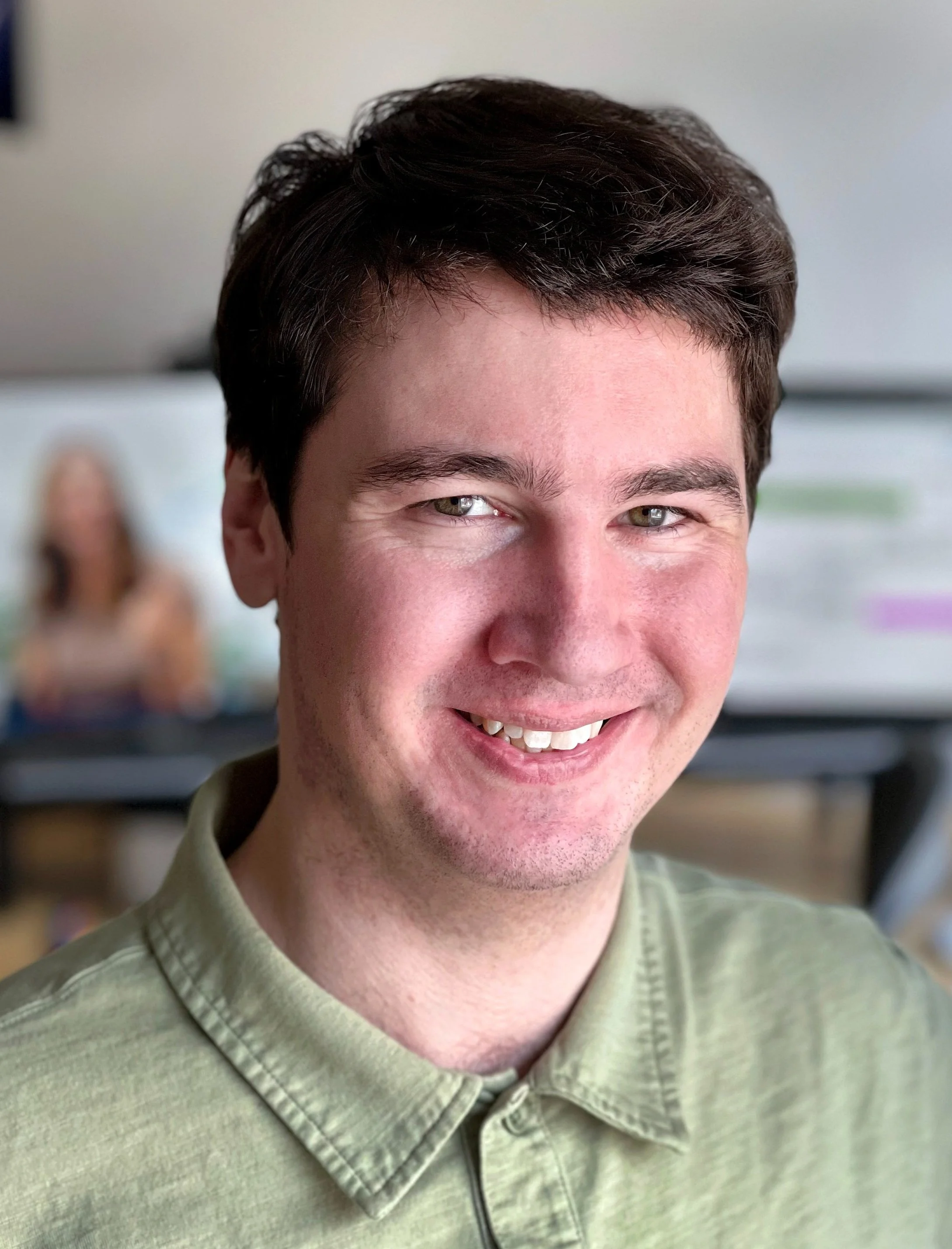 A man smiling, wearing a green collared shirt, with a blurred background.