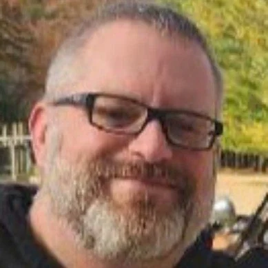 A bearded man wearing glasses, smiling outdoors with blurred trees in the background.