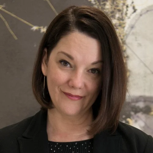 Portrait of a woman with dark shoulder-length hair, wearing a black blazer and earrings, set against a neutral artistic background.