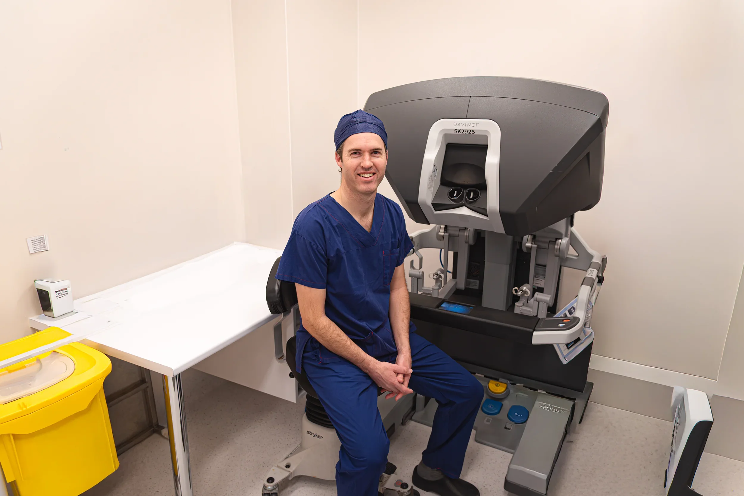 A medical professional in blue scrubs and a surgical cap sitting in a hospital room. He is smiling, and behind him is a large medical imaging machine, likely an MRI or CT scanner.