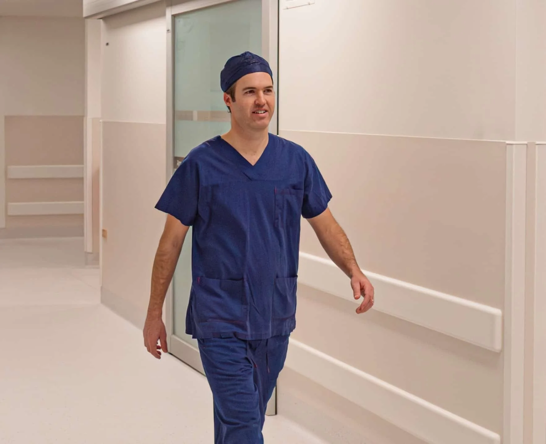 A male healthcare worker in blue scrubs and a cap walking down a hospital corridor.