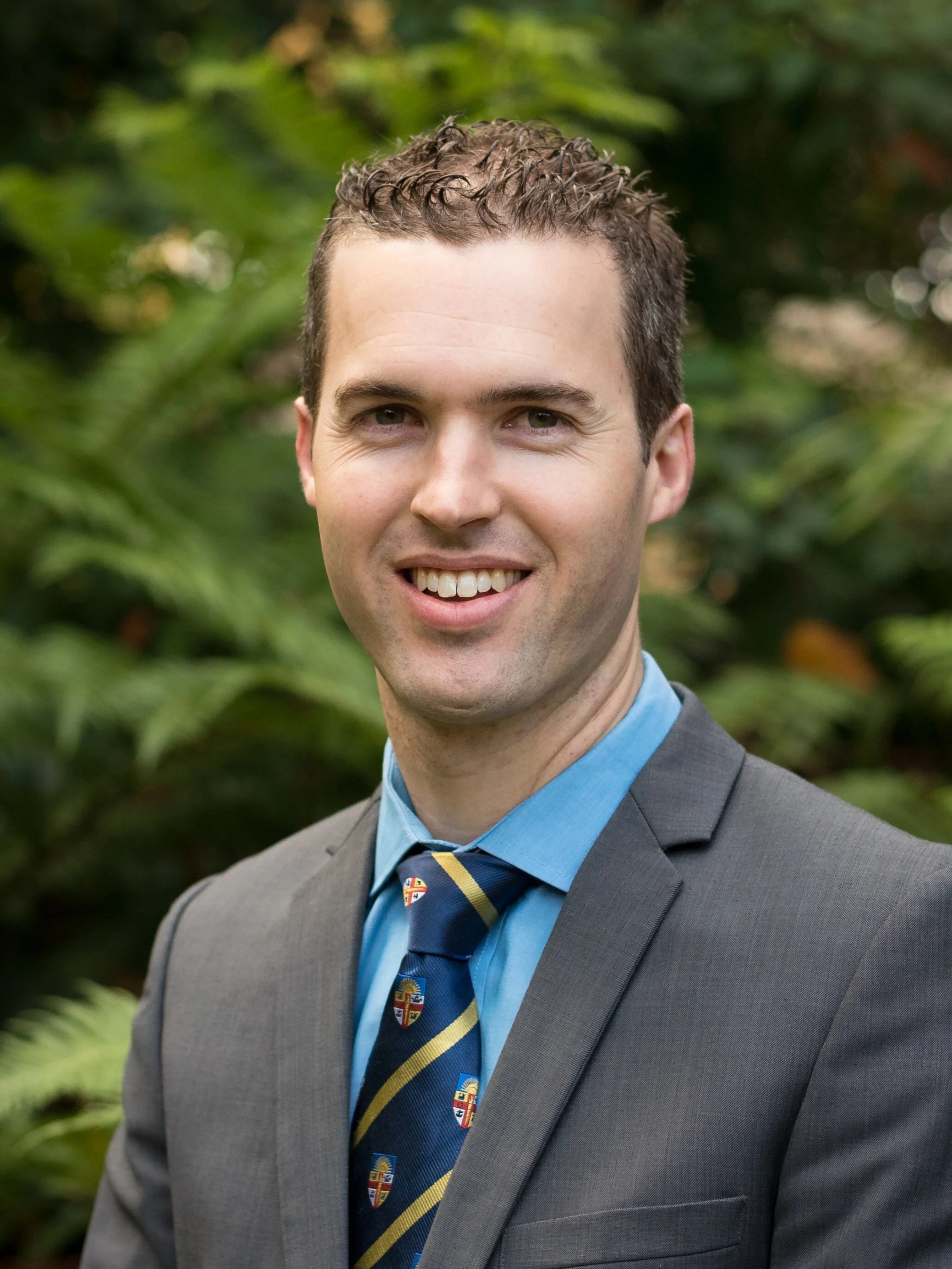 A young man with short, curly brown hair smiling in a business suit and tie, standing outdoors with blurred green foliage in the background.