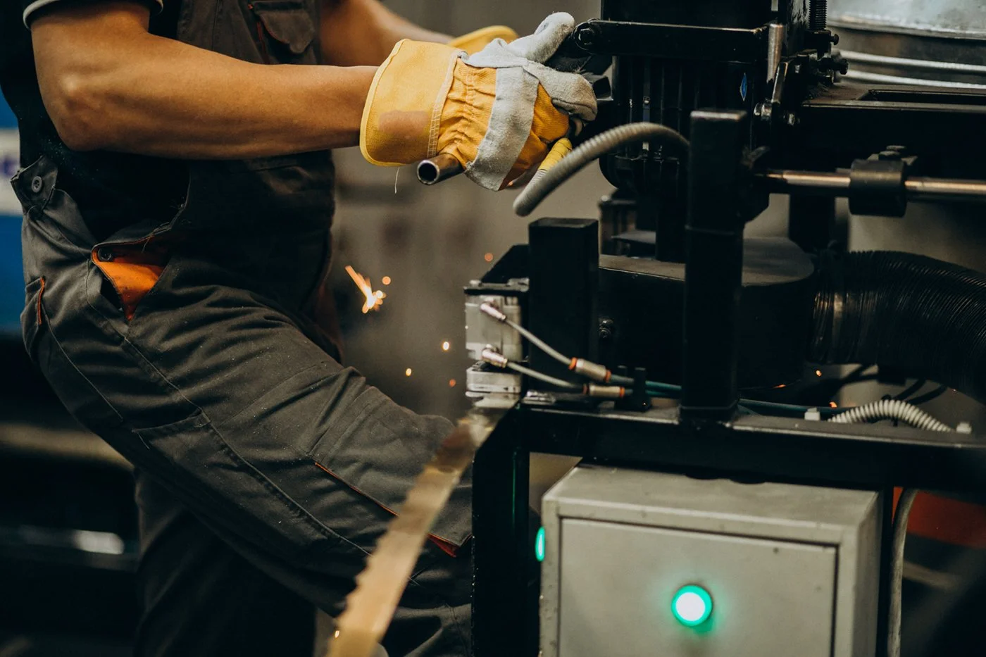 A worker welding metal in an industrial setting, wearing protective gloves. Sparks are flying as they weld a metal piece to a machine.