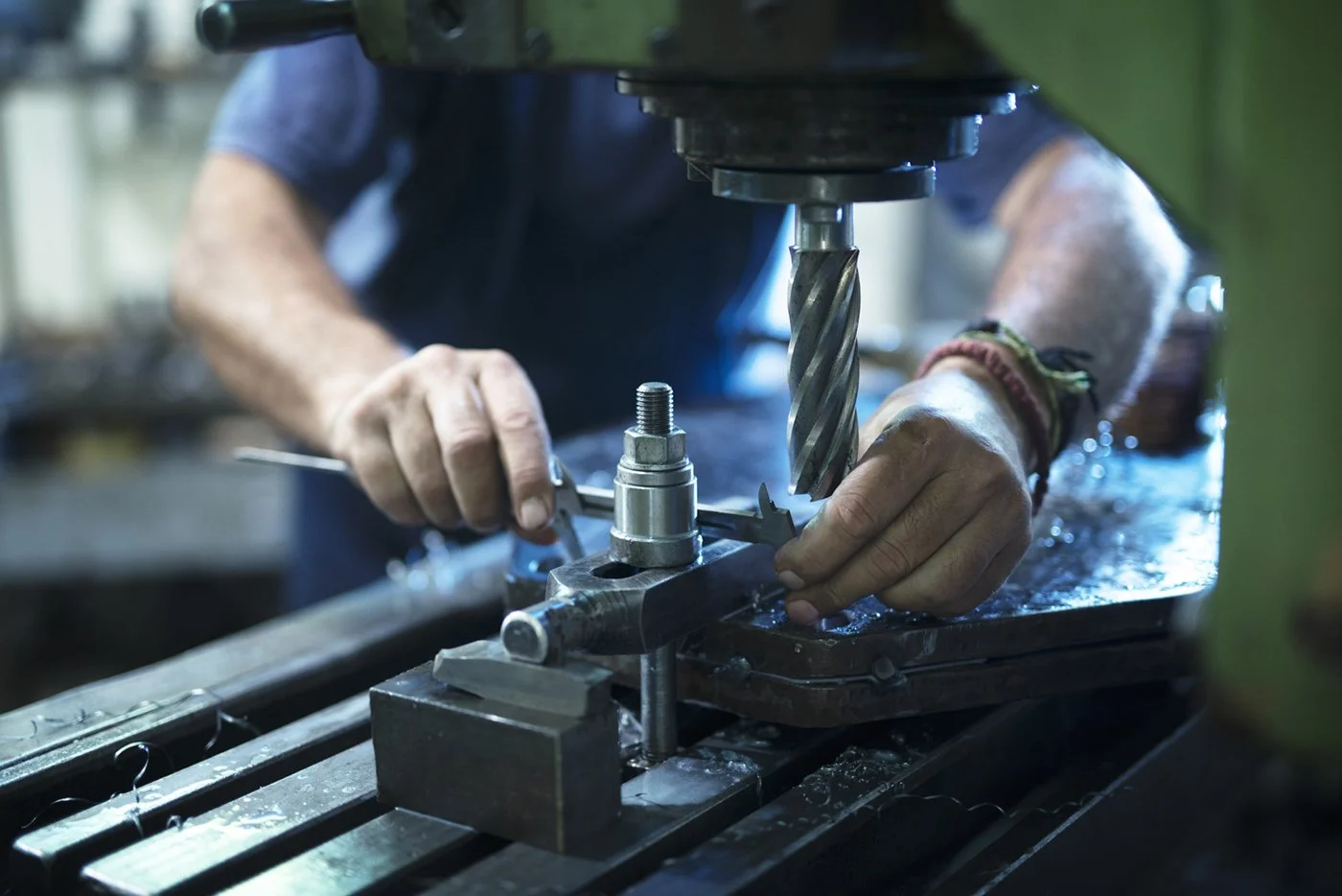 A person operating a drill press in a workshop, using a caliper to measure a metal component.