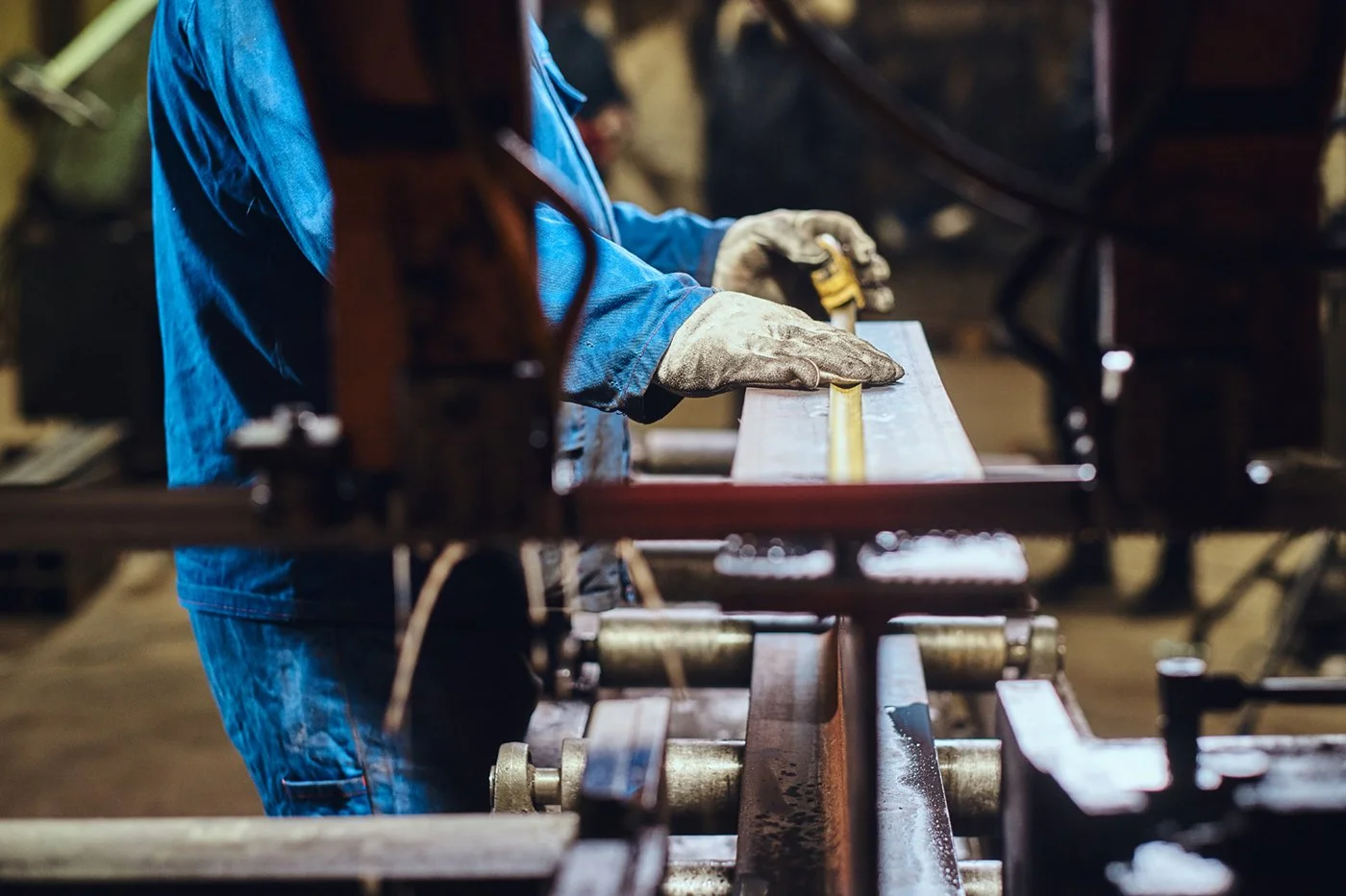 A worker in a blue jumpsuit and gloves is using a measuring tape on a piece of metal in a workshop.