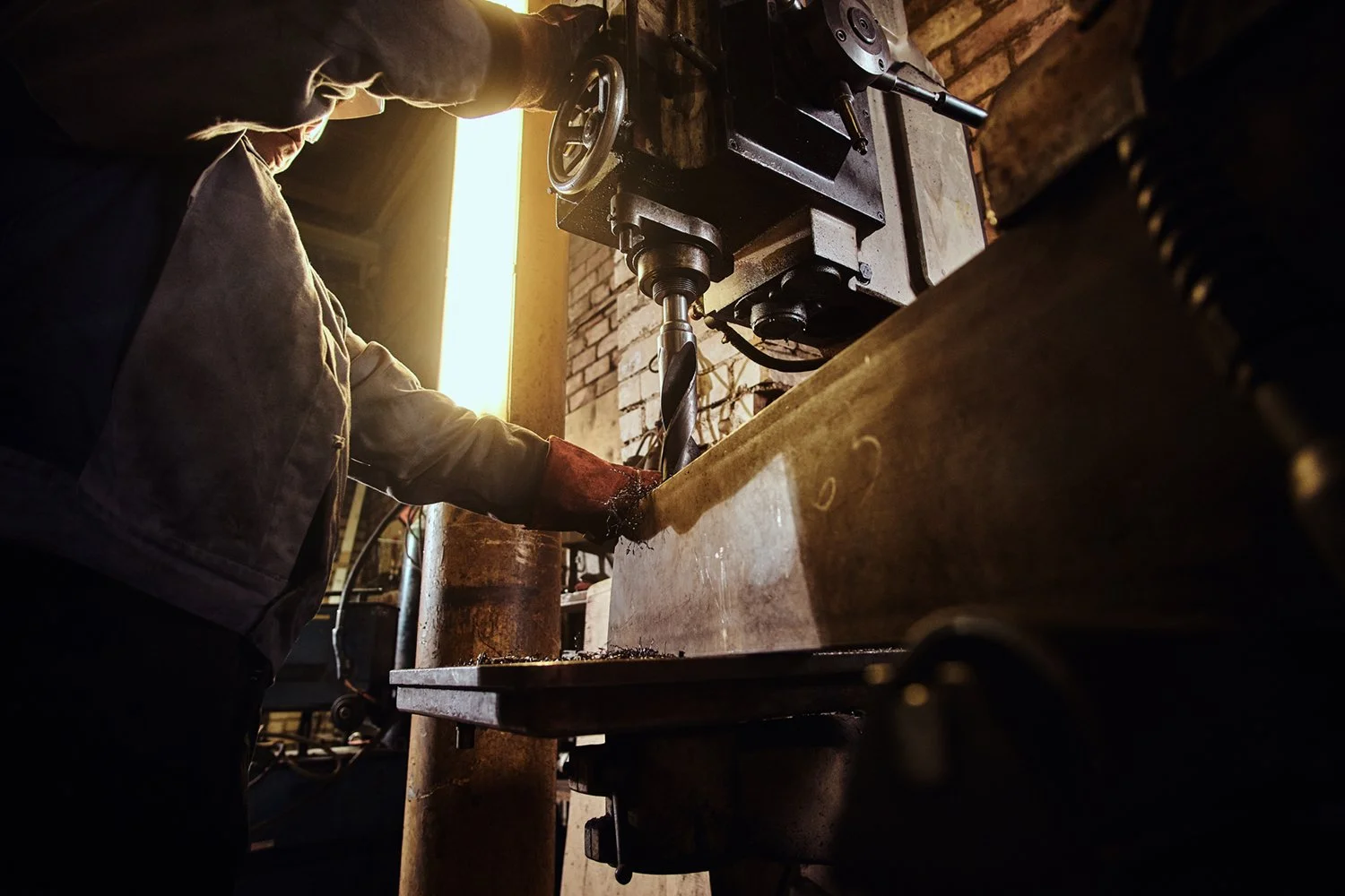 A person working in a workshop operating a large industrial drill press, with a brick wall in the background and warm lighting.