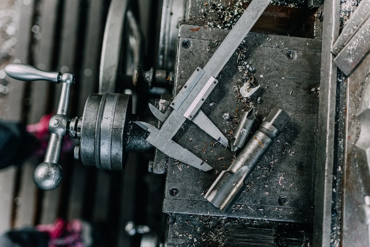 Close-up of a metal lathe machine with a metal part and measurement tools on a workbench, surrounded by metal shavings.