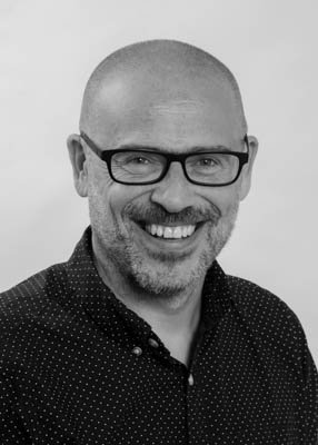 Black and white photo of a smiling man with glasses, a beard, and a shaved head, wearing a collared shirt.