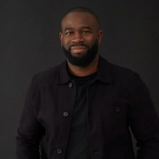 A man with a beard wearing a black jacket and black shirt, standing against a dark background.