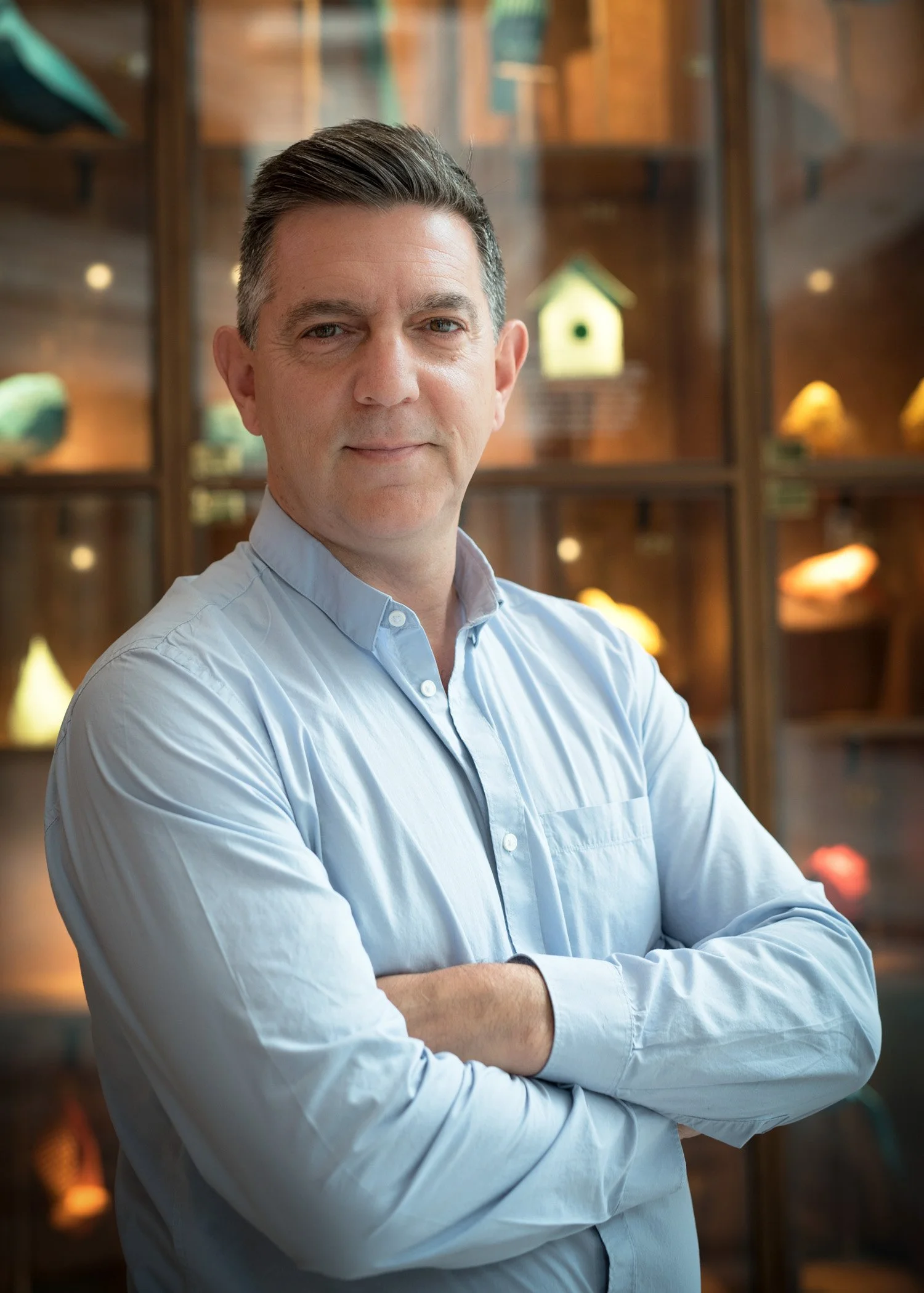 A man with short dark hair, wearing a light blue button-up shirt, standing with arms crossed in front of a wooden display case with various objects.