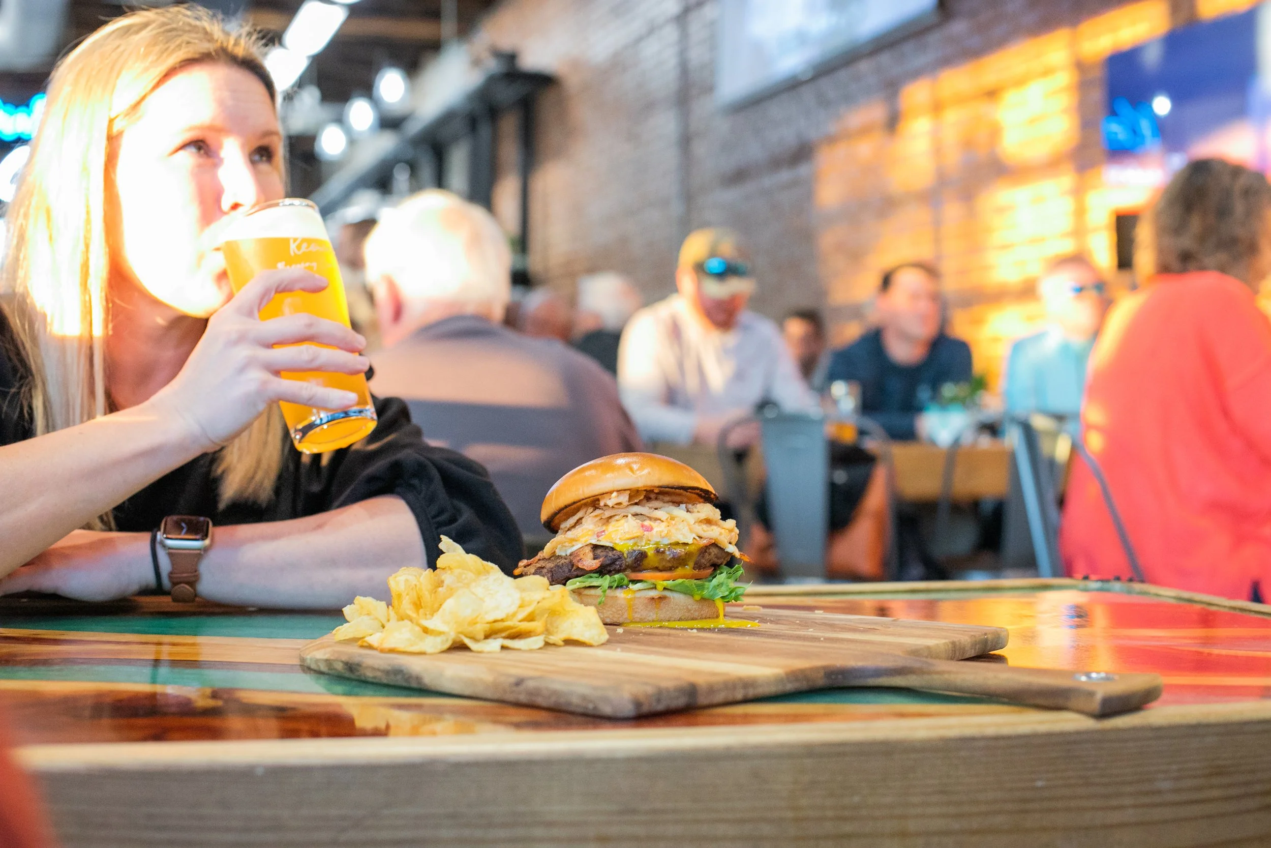 Young lady sipping a beer alongside a large juicy burger and chips.