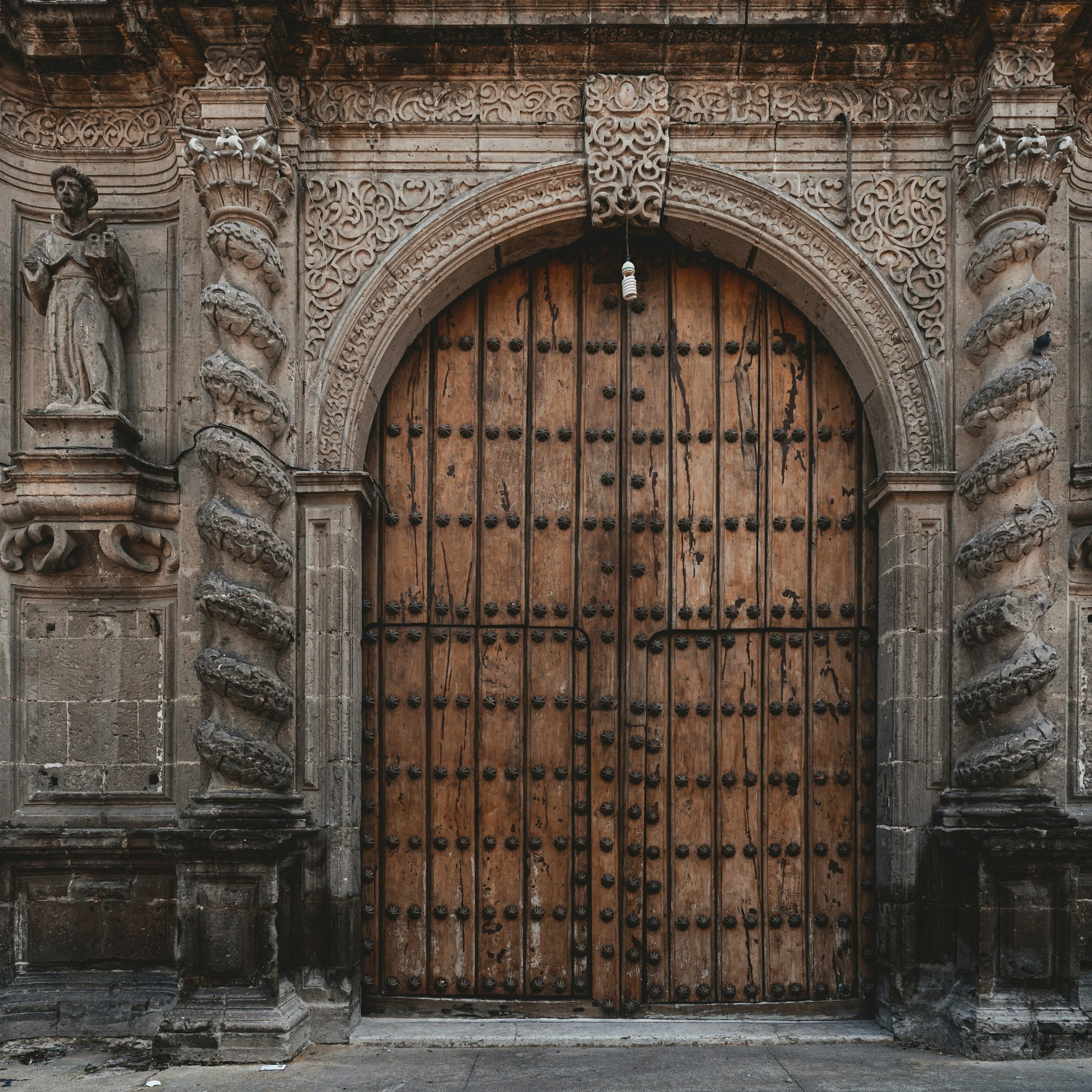 Large wooden church door with decorative iron studs inside an ornate stone archway with carved statues on the sides.