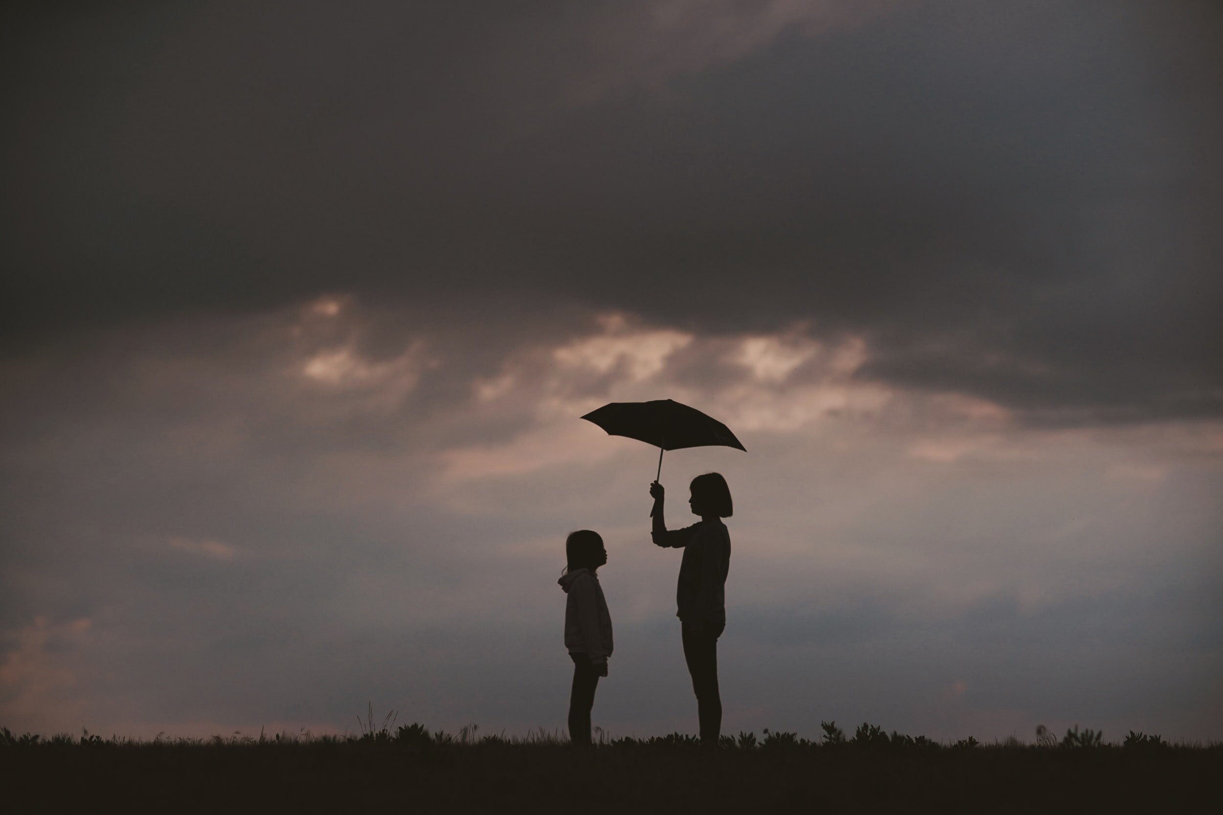  An adult stands in the rain as they hold an umbrella above their head. They are also providing shelter for a younger child. This could symbolize what it is like to have support from a trauma therapist in Dallas, TX. PTSD treatment could help you ove