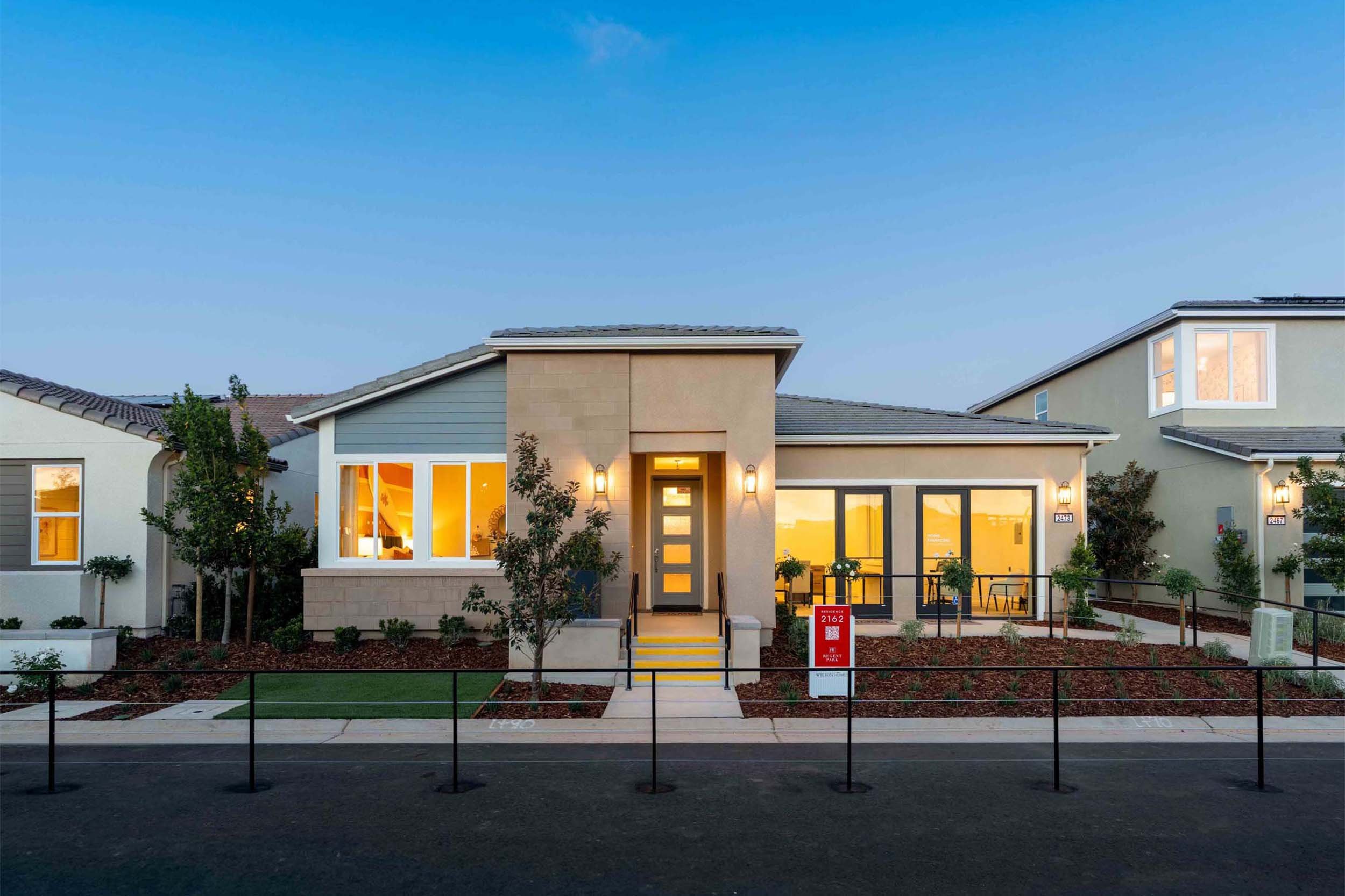 Contemporary house with large windows and front porch, illuminated from inside, surrounded by a landscaped garden with small trees, plants, and a sidewalk, during twilight or early evening.