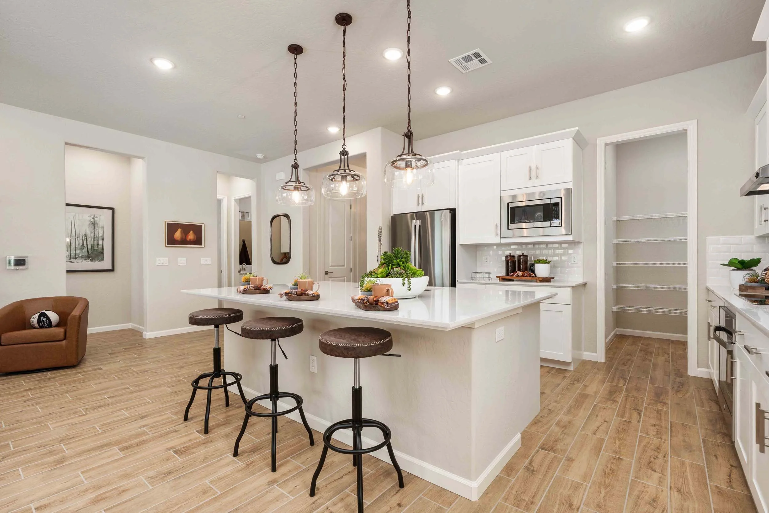 A modern kitchen with white cabinets, stainless steel refrigerator and microwave, a large white island with a plant and tableware, three brown bar stools, hanging pendant lights, and a wooden floor.