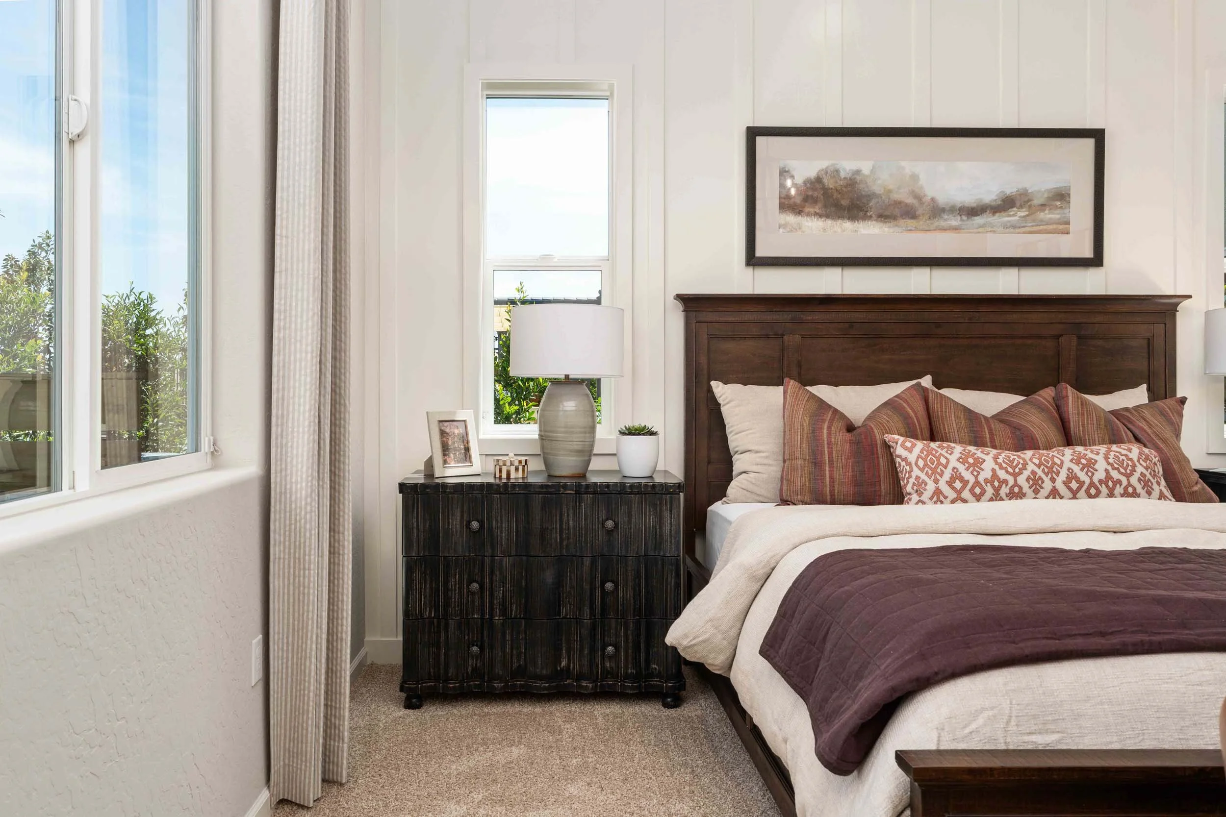 A bedroom with a wooden bed frame, patterned pillows, and a quilt. A nightstand with a lamp, a framed photo, and plants. Two windows with curtains, and a framed landscape picture on the wall.