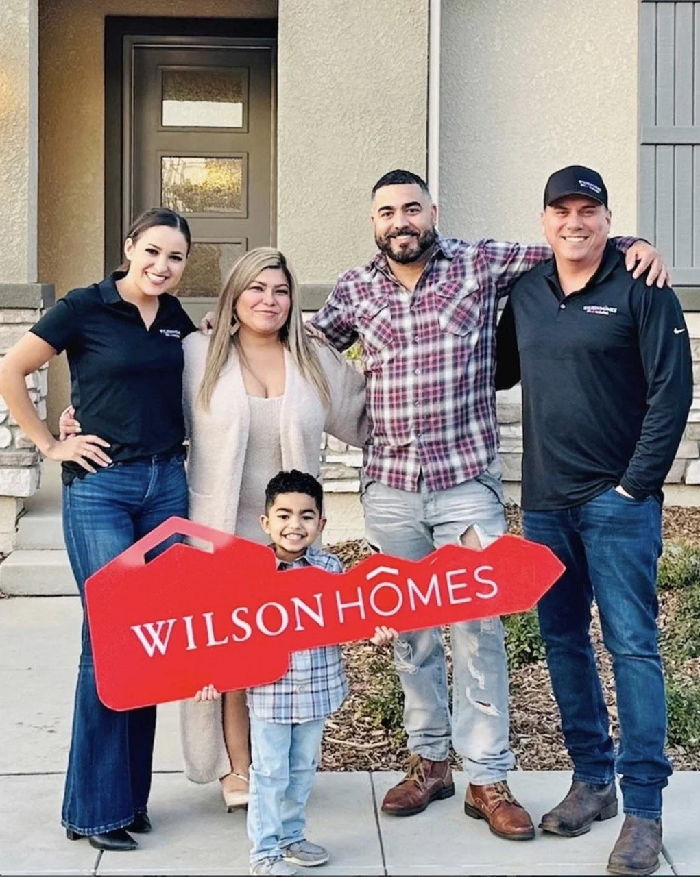 A group photo of five people standing in front of a house with a modern door. They are smiling, and the group includes a young boy holding a red sign that reads "Wilson Homes." The others are adults, and they appear happy, celebrating in front of the home.