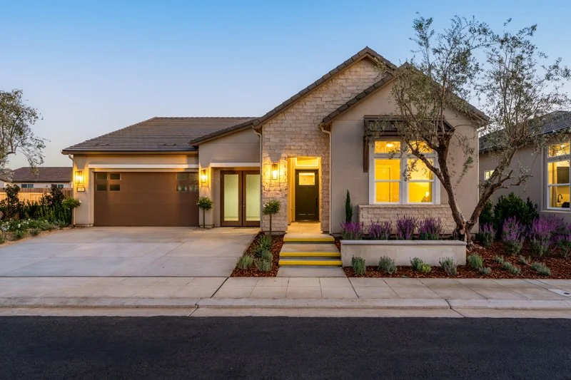 Modern suburban house with a two-car garage, front steps, illuminated entryway, landscaped yard with trees and purple flowers, and warmly lit windows during dusk.
