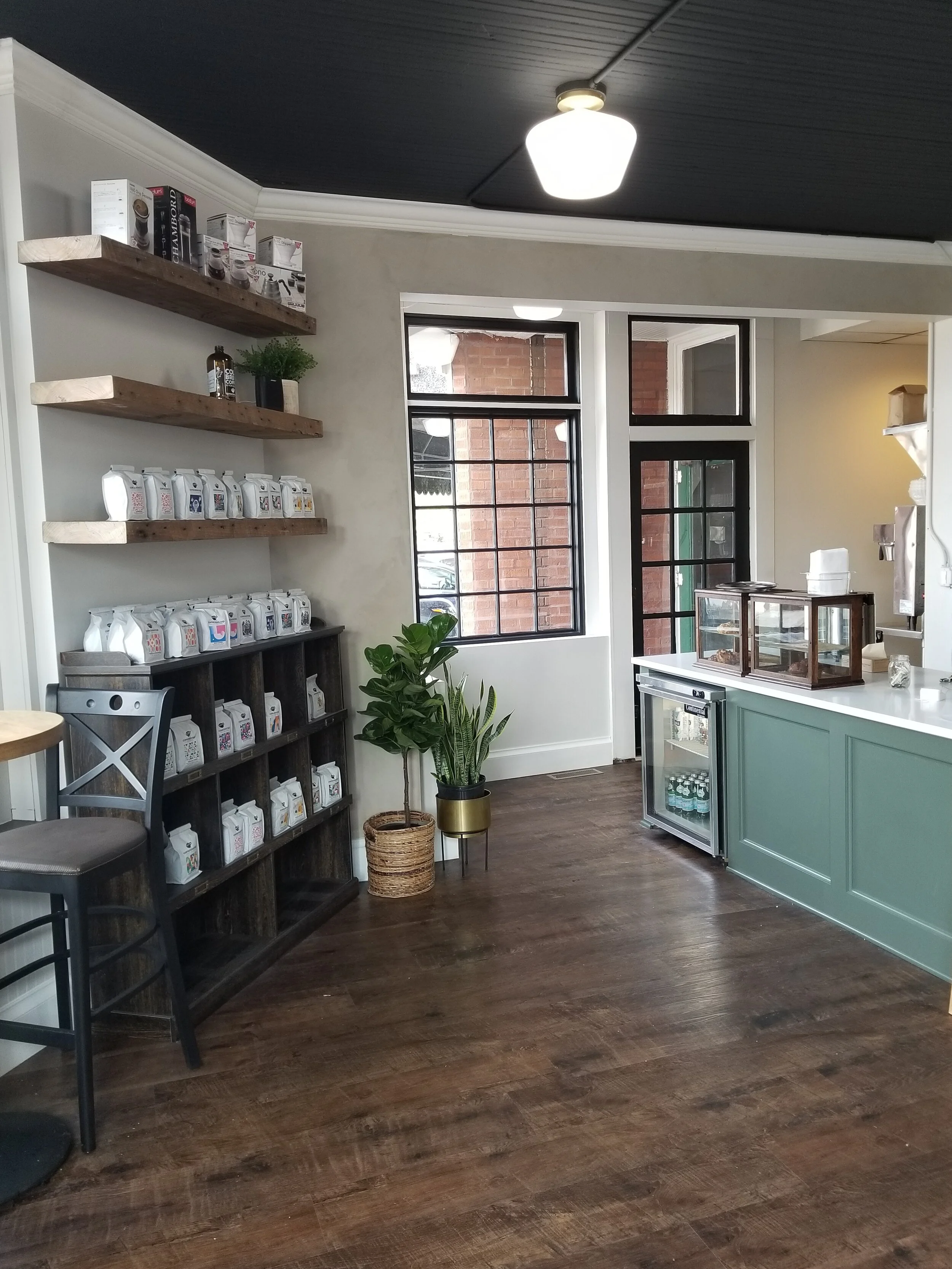 Interior of a coffee shop with wooden floors, black ceiling, white walls, and shelves holding coffee bags and products. A small refrigerator with beverages, green counter, and plant in a pot are visible near the window.