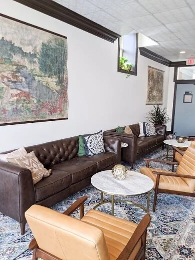 A cozy waiting area with a long brown leather sofa, colorful pillows, and two beige armchairs around a round marble coffee table with a decorative orb. The room has a patterned rug, framed artwork on white walls, and a small window with a potted plant.