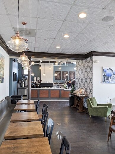 Interior of a cozy cafe with wooden tables and chairs, a green armchair, and decorative wallpaper on one wall, with a counter area in the background.