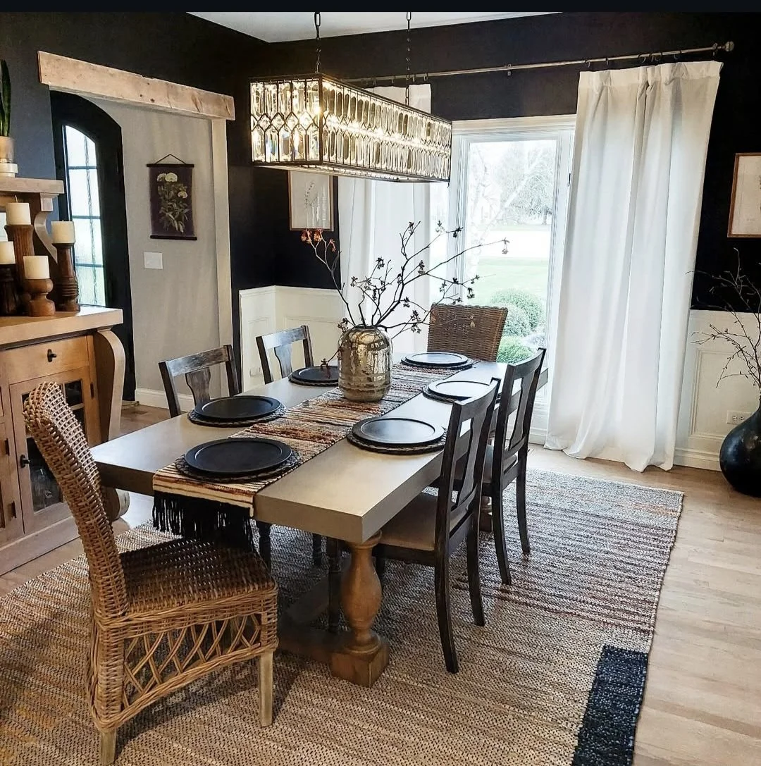 A dining room with a wooden table set for five, featuring dark plates, a woven table runner, and a large vase with bare branches as a centerpiece. The room has black and white walls, white curtains, a large window/door with a view outside, and a rectangular chandelier overhead. There is a woven chair at one end of the table and a wooden sideboard with candles nearby.
