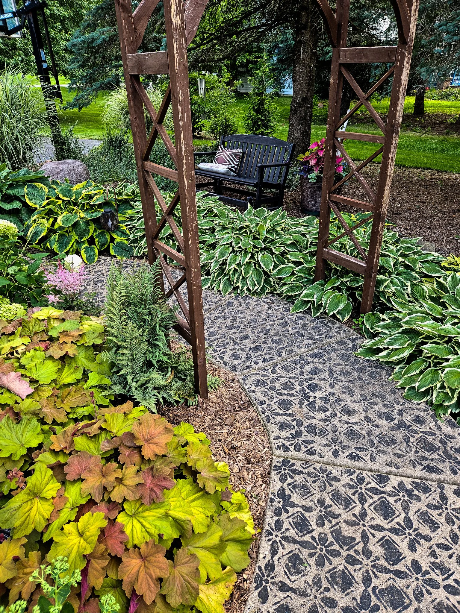 A garden scene featuring a stone pathway, lush green and variegated plants, a wooden arch, a black garden bench, and potted pink flowers in the background.
