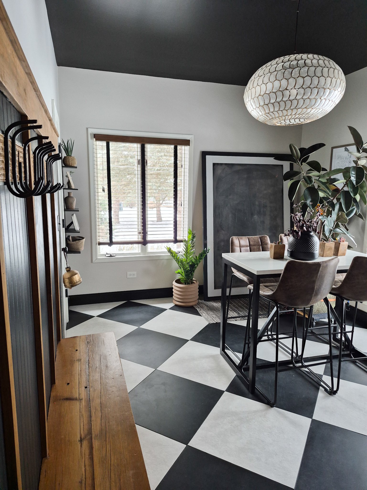 Modern dining area with black and white checkered floor, a white table, and beige chairs. Features a large blackboard, a potted plant near a window, and a decorative ceiling light.