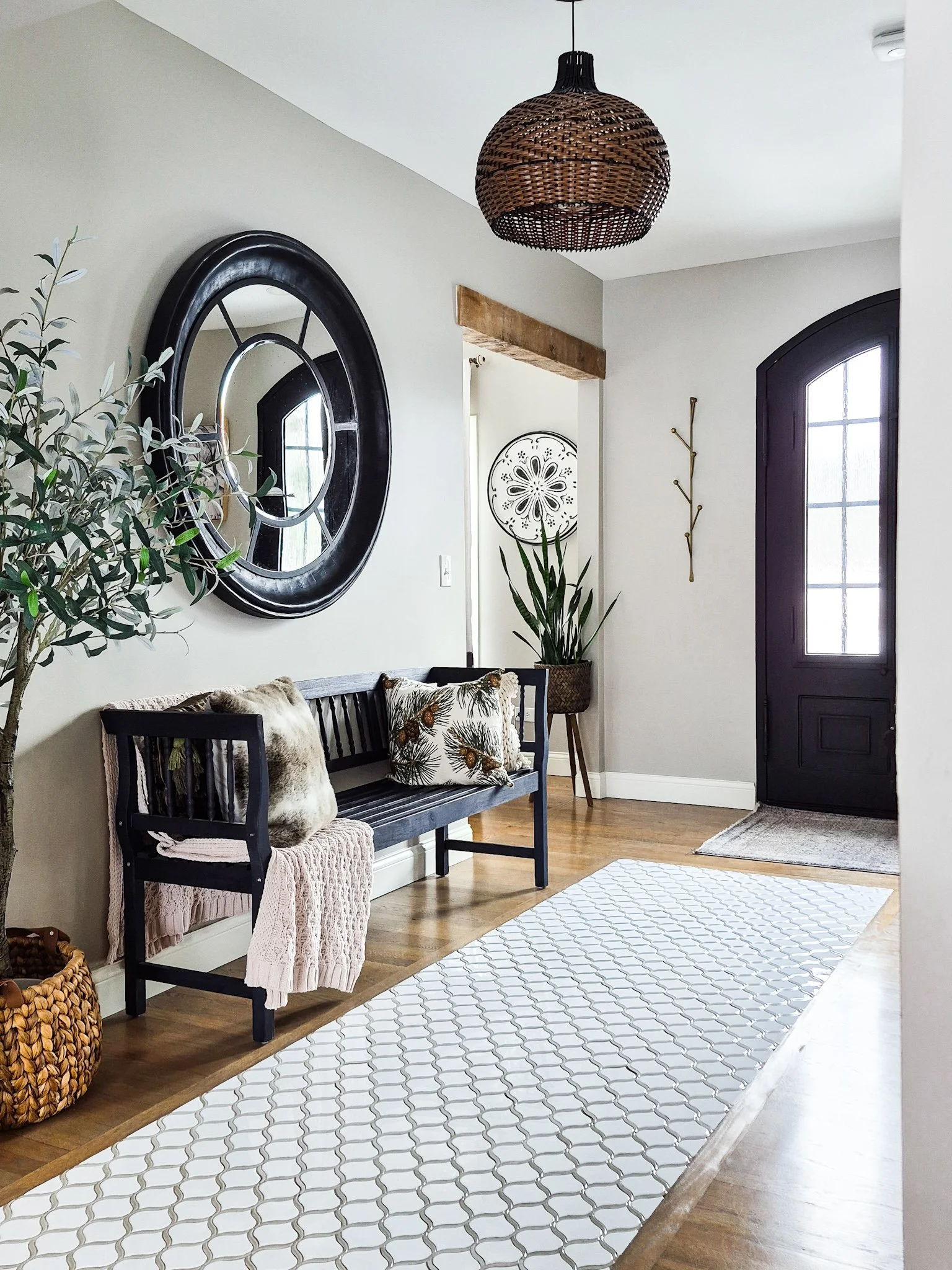 Entryway with wooden floor, black front door with glass panels, white walls, a black bench with cushions and throw blankets, a large round mirror, a potted plant, a woven basket, and decorative wall art.