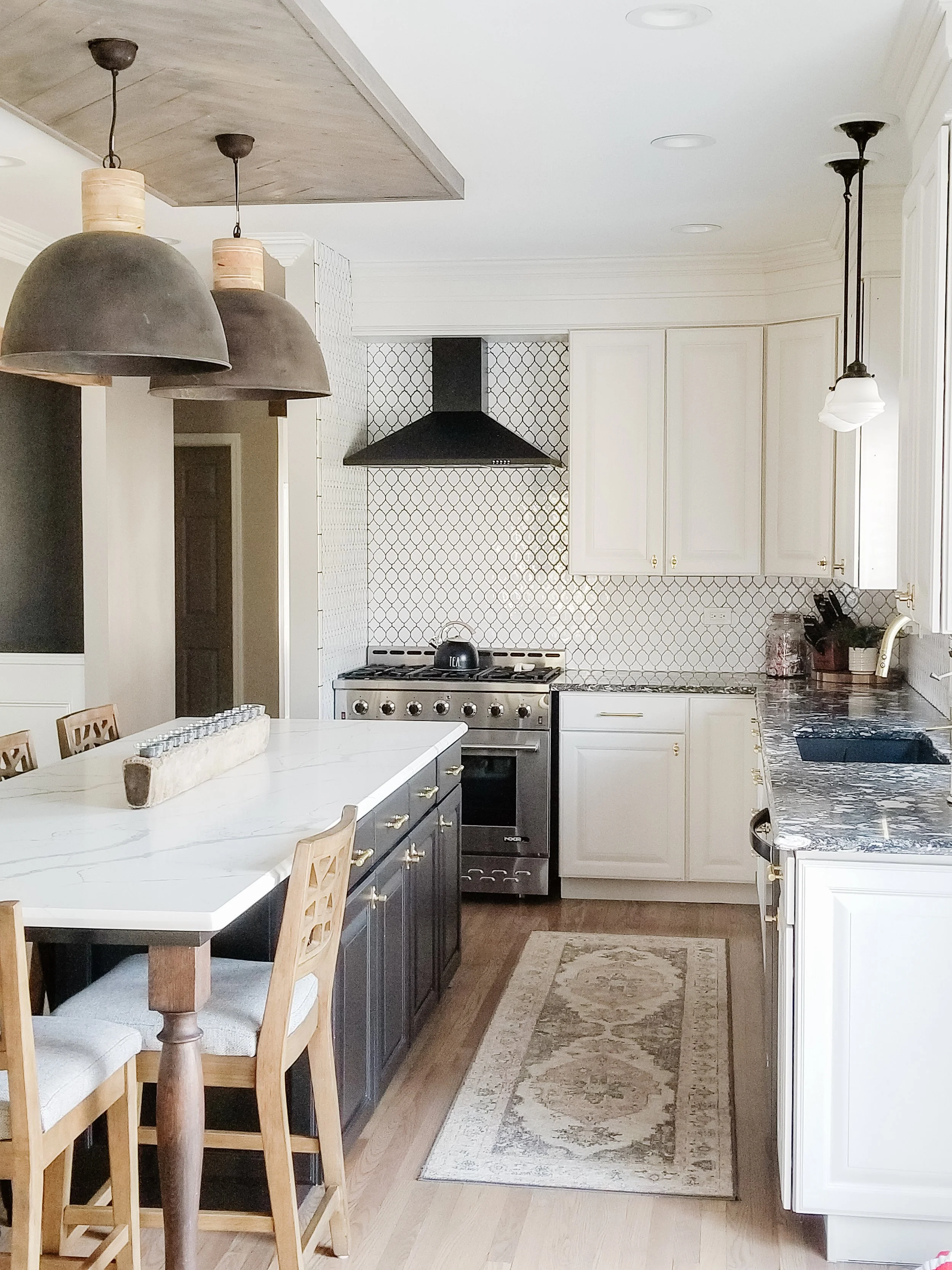 A kitchen with white upper cabinets, a black range hood above a stove, a marble island with wooden chairs, patterned backsplash, and hanging pendant lights.