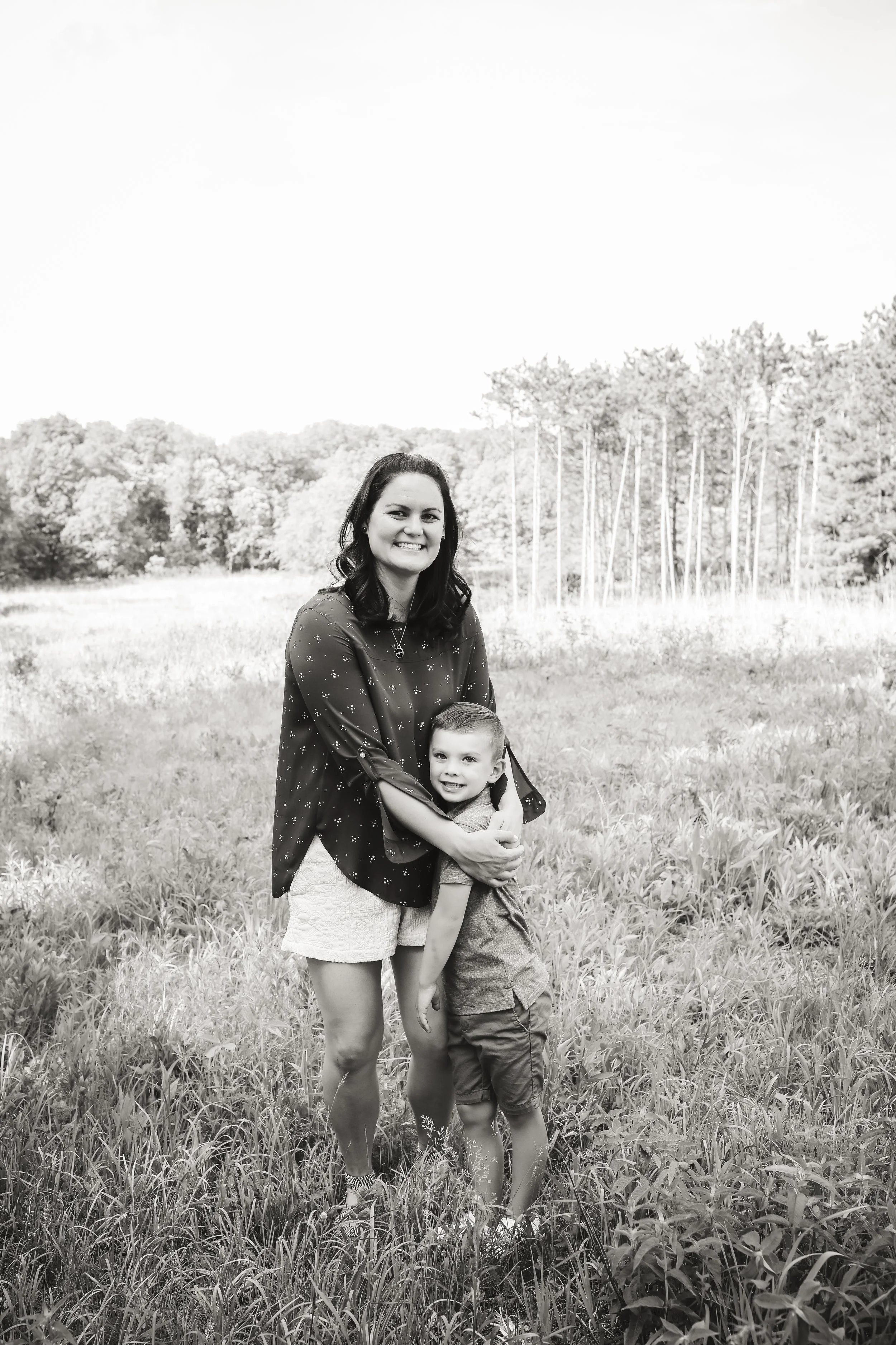 A woman and a young boy standing in a grassy field, smiling. The woman has dark hair and is wearing a patterned blouse and shorts, and the boy is wearing a T-shirt and shorts. They are hugging and enjoying a day outdoors with trees in the background.