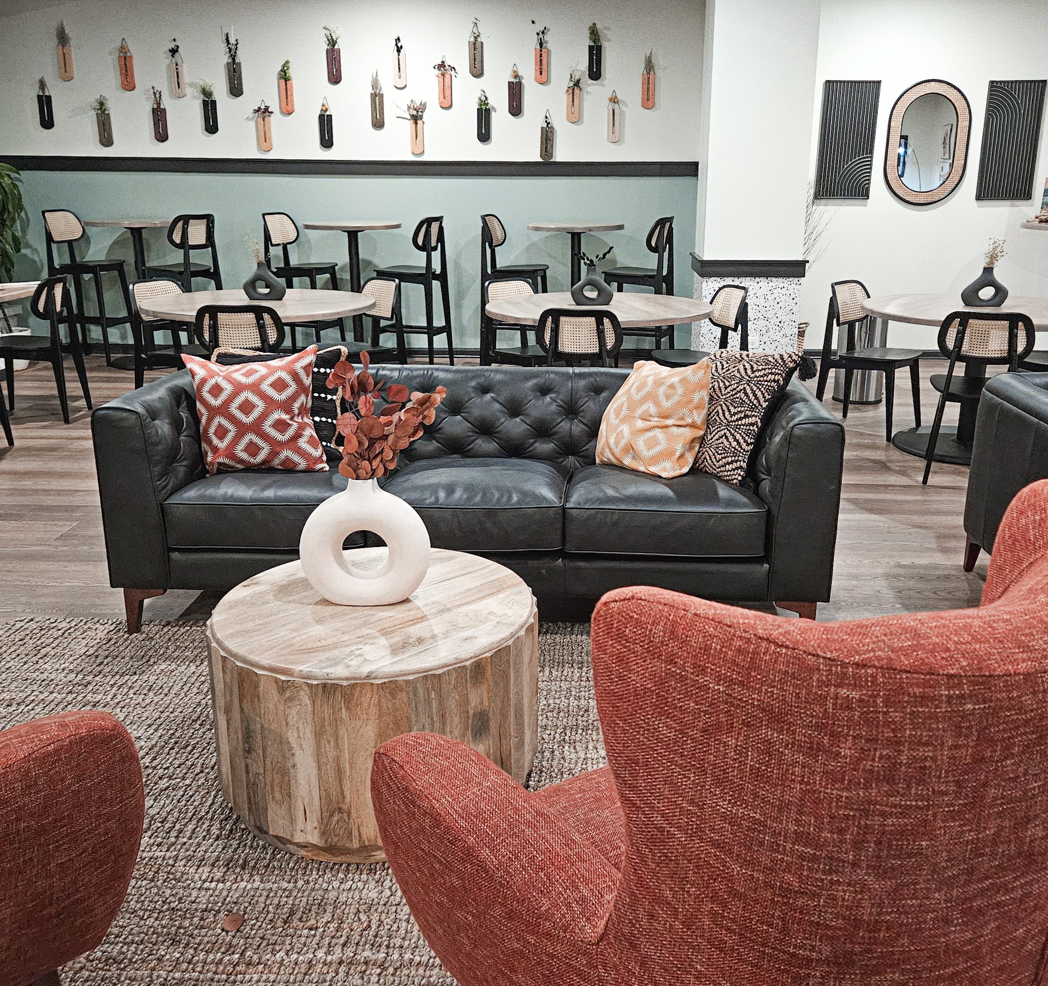 A cozy lounge area featuring a black leather tufted sofa with three decorative pillows, a round wooden coffee table with a white ceramic vase holding dried red leaves, red armchairs, a patterned rug, and a background wall decorated with small framed plants and hanging wall art. There are additional tables and chairs along the wall, with a large mirror and artwork on the white wall.