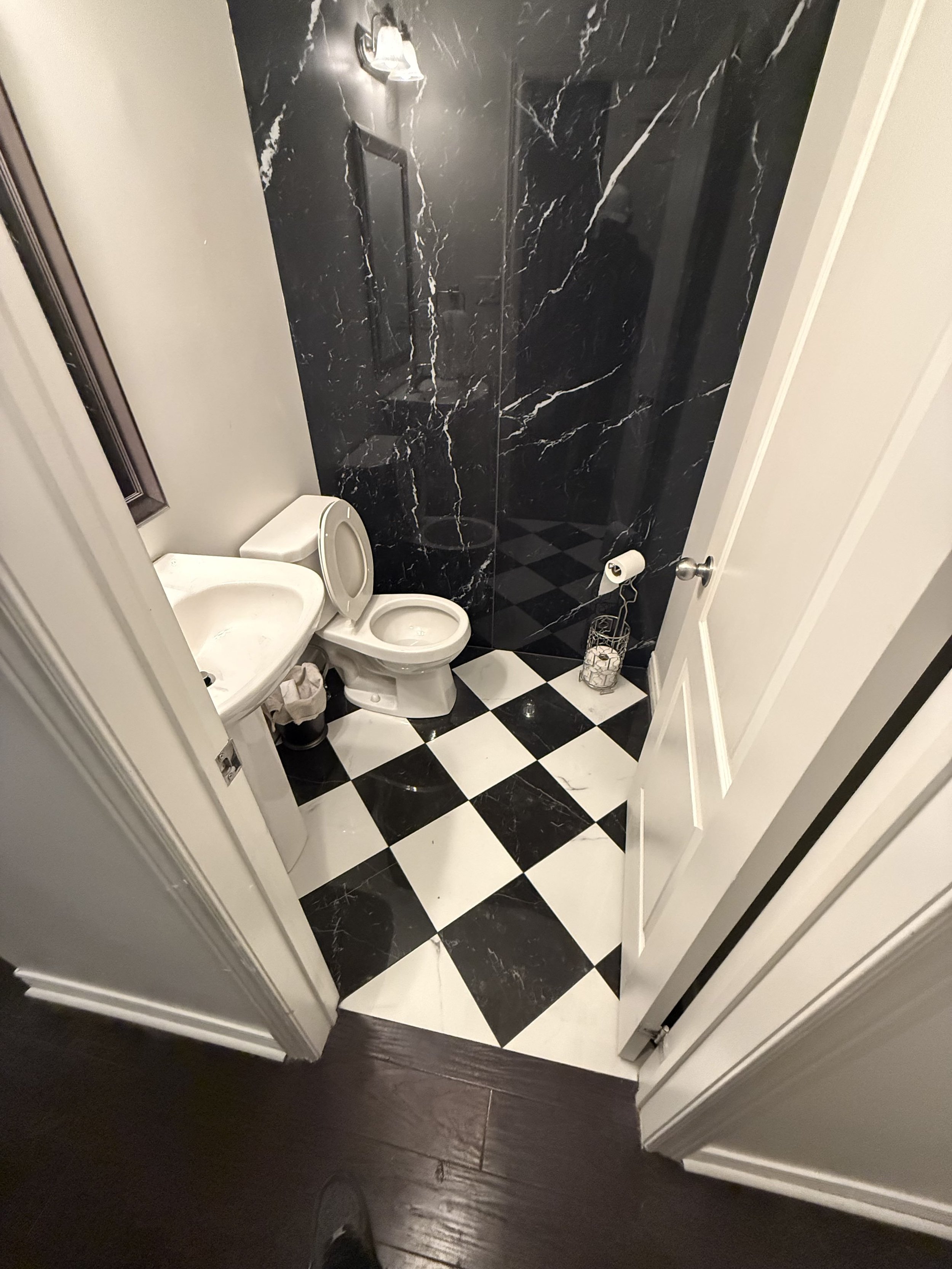 A small bathroom with black and white checkered flooring, a toilet, a small sink, and a black marble shower area. There is a wall-mounted light and a mirror above the sink.